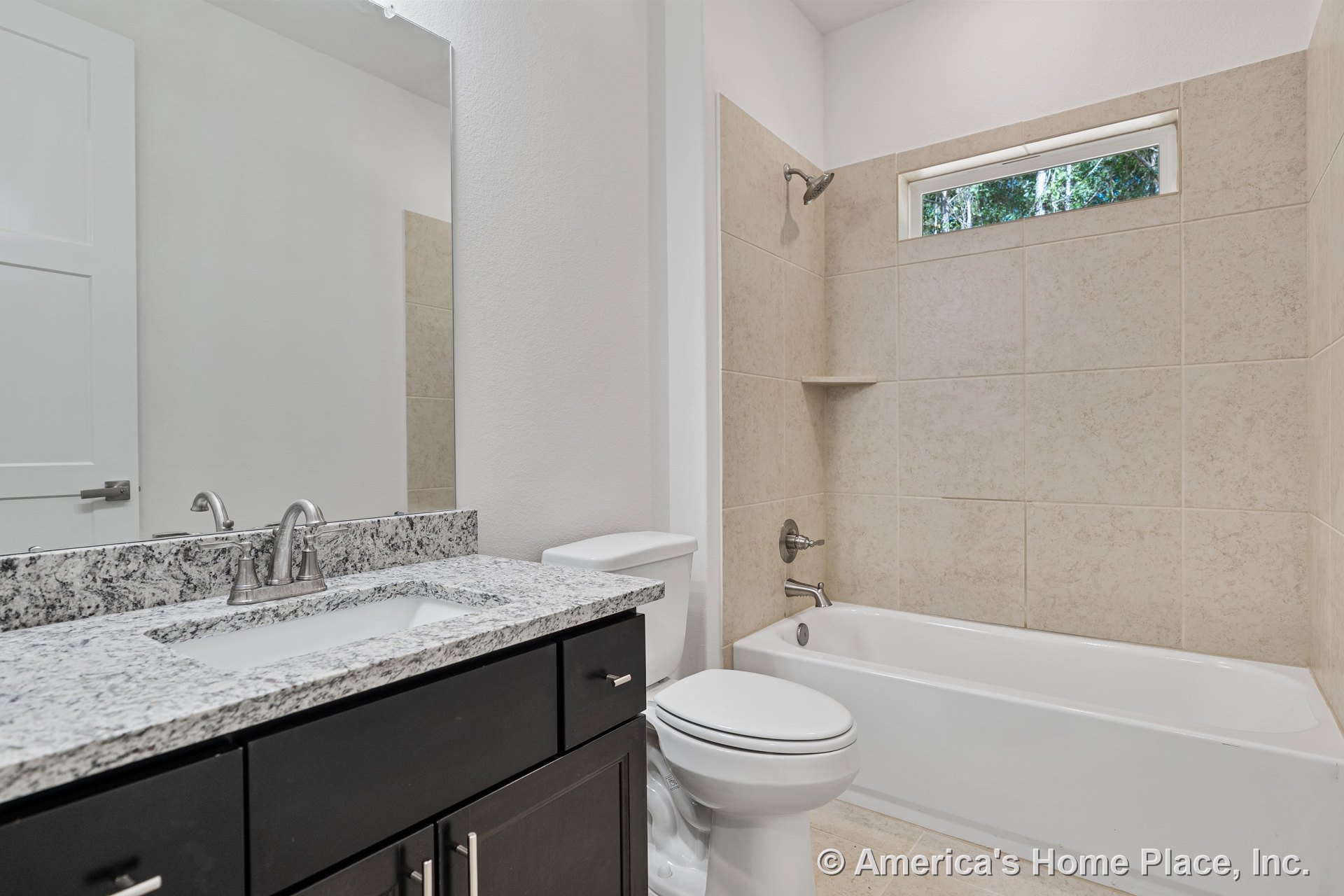 Granite countertop atop dark wood vanity, large wall mirror, tiled tub-shower combo with built-in shelf, high horizontal window, chrome fixtures, and floor tile.
