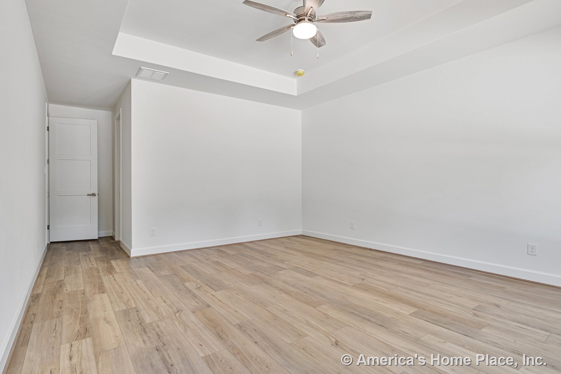 Bedroom with light wood plank flooring, tray ceiling featuring a modern ceiling fan, white trim and baseboards, three-panel interior door, and neutral wall color.