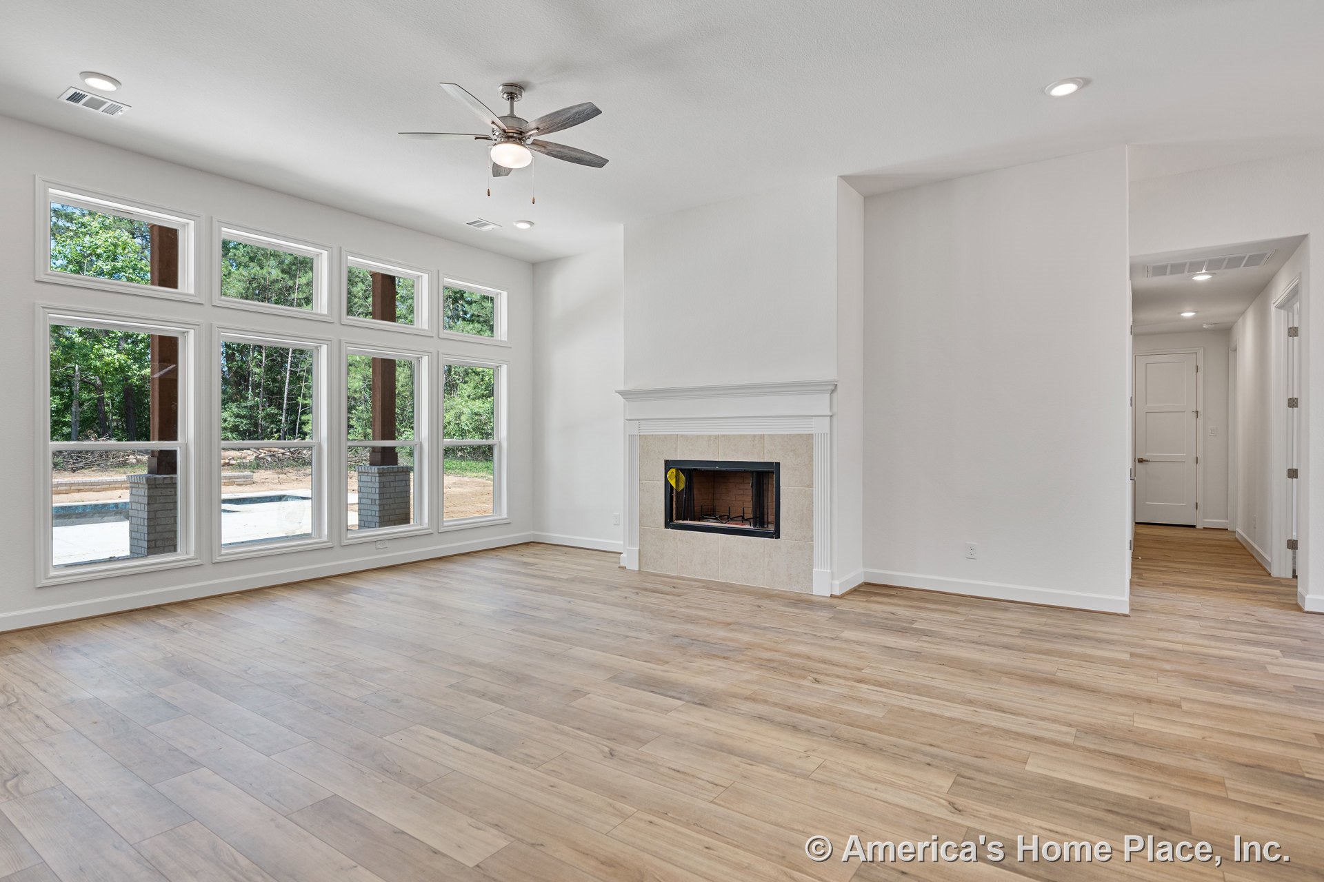 Tiled fireplace with white mantel, expansive grid windows, light wood plank flooring, ceiling fan with integrated light, recessed ceiling lights, white interior trim, and open