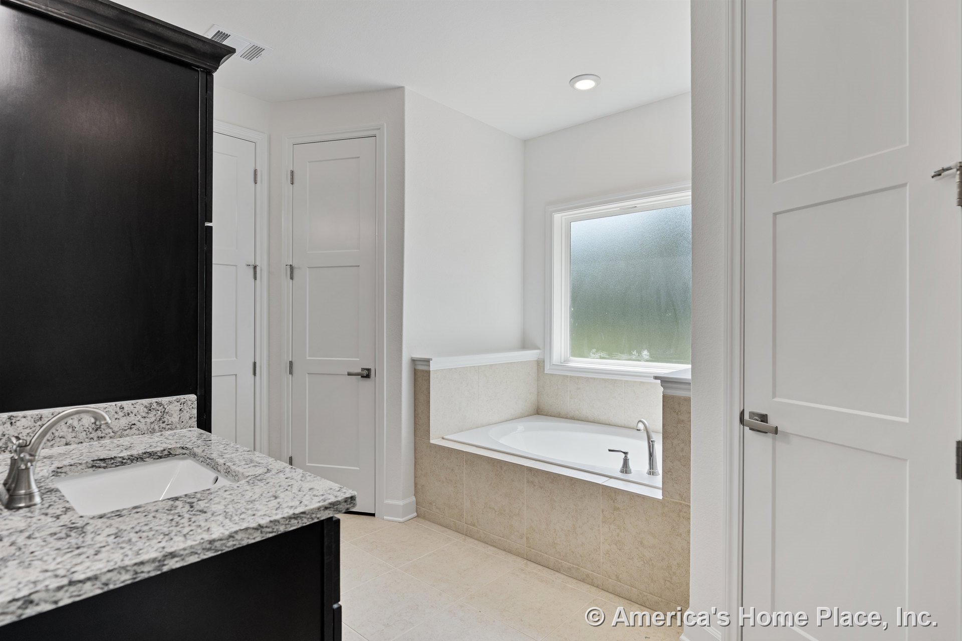 Built-in soaking tub with tile surround beneath a frosted window, granite countertop vanity, recessed ceiling light, white paneled doors, and neutral tile flooring.