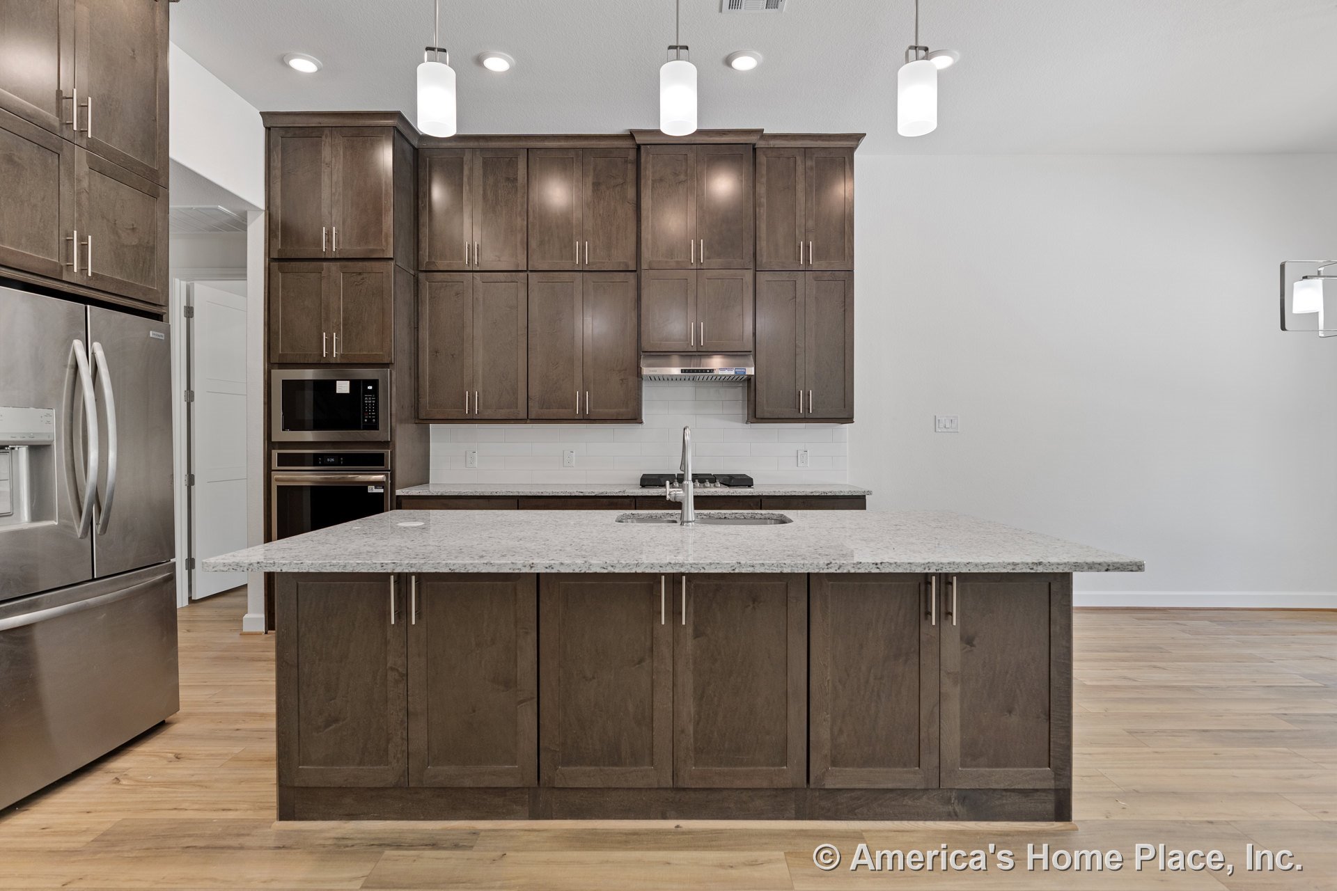 Dark wood cabinetry kitchen with large granite island countertop, stainless steel refrigerator, built-in microwave and oven, gas cooktop, white tile backsplash, pendant and