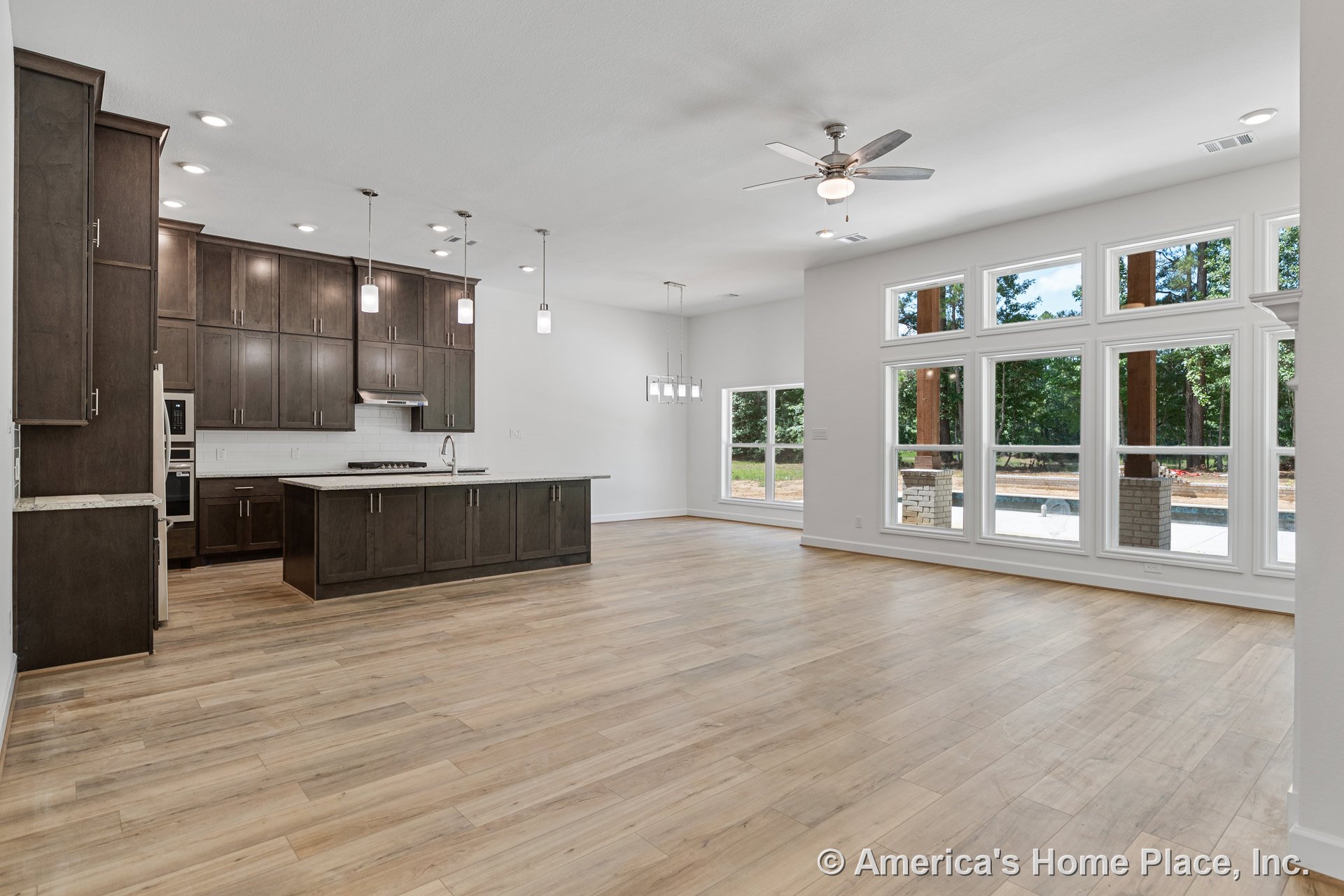 Dark wood kitchen cabinetry and large island with quartz countertops, pendant and recessed ceiling lights, wide plank wood flooring, multiple tall windows with white trim, ceiling