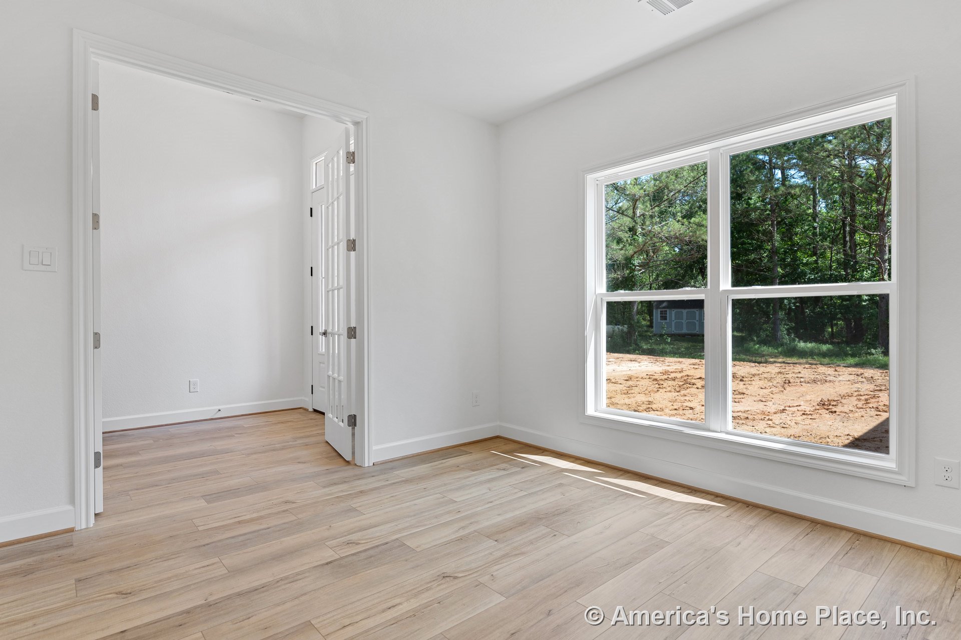Large double windows with white trim fill the room with natural light, highlighting light wood-look tile flooring, white walls, baseboards, an open doorway with white casing, and a