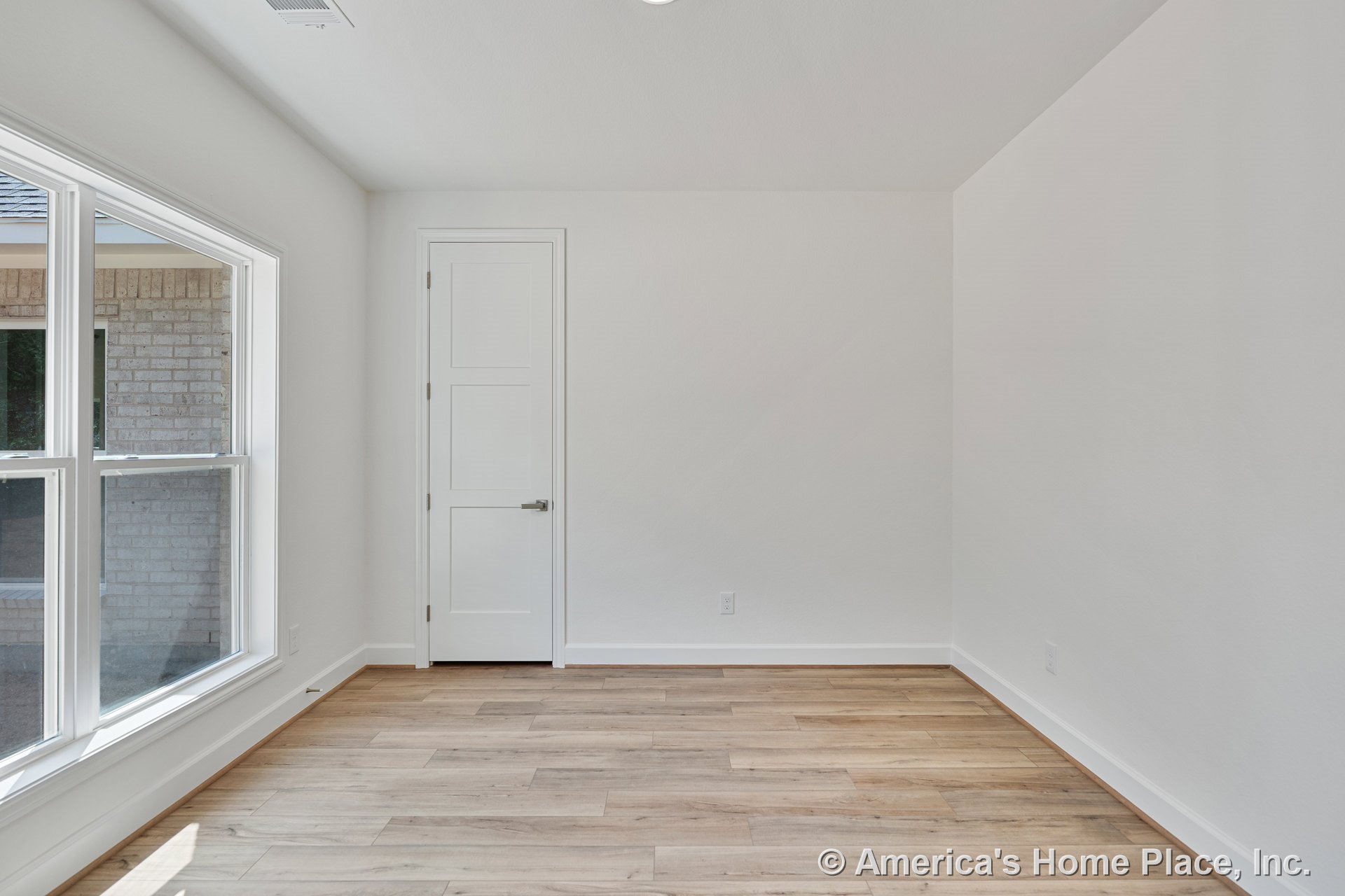 Large double windows with white trim fill the room with natural light, highlighting light wood plank flooring, white walls, baseboard molding, and a three-panel interior door