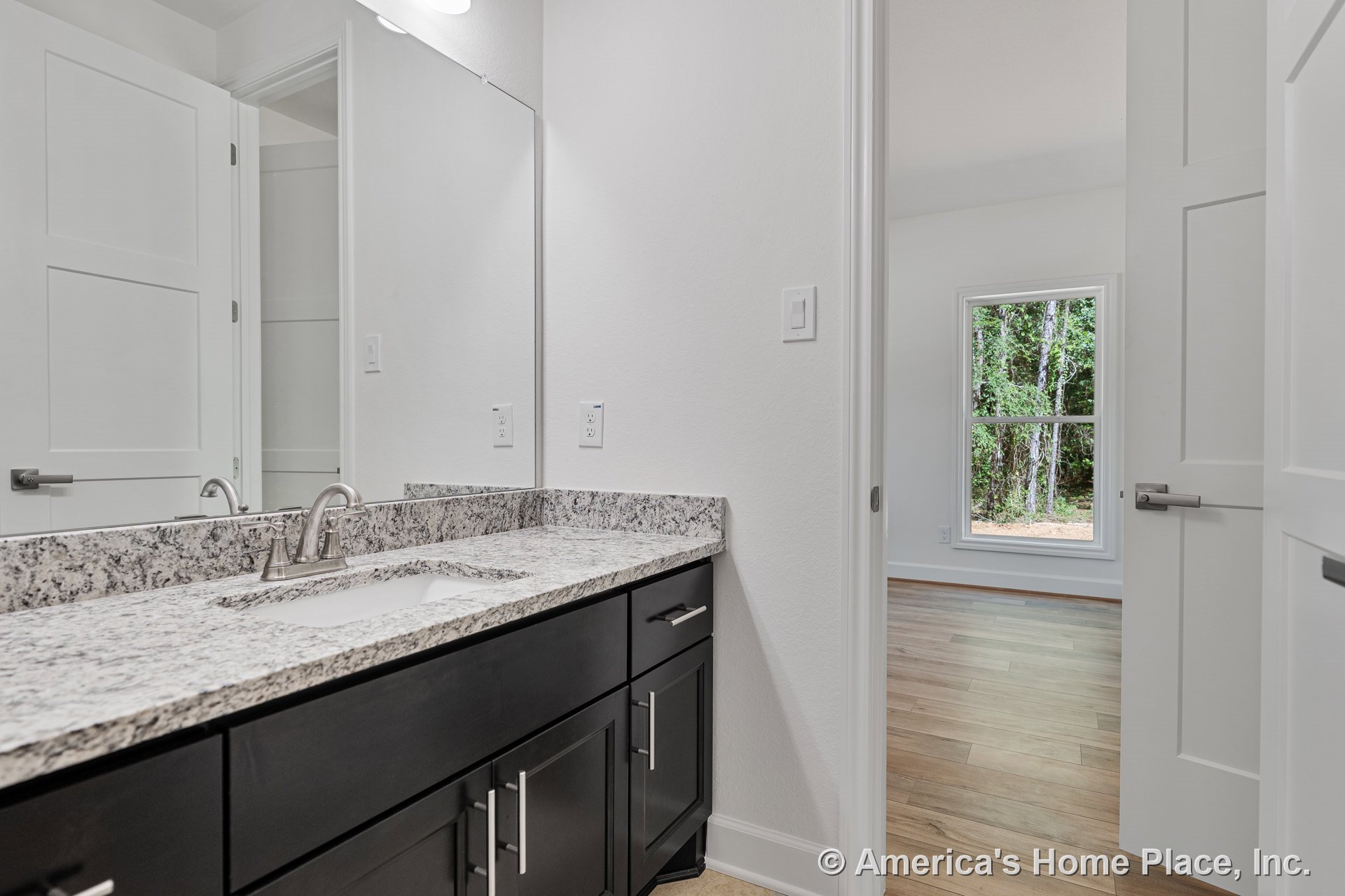Granite bathroom countertop with undermount sink and brushed nickel faucet atop dark wood vanity cabinets, large wall mirror above, white paneled doors leading to bedroom with