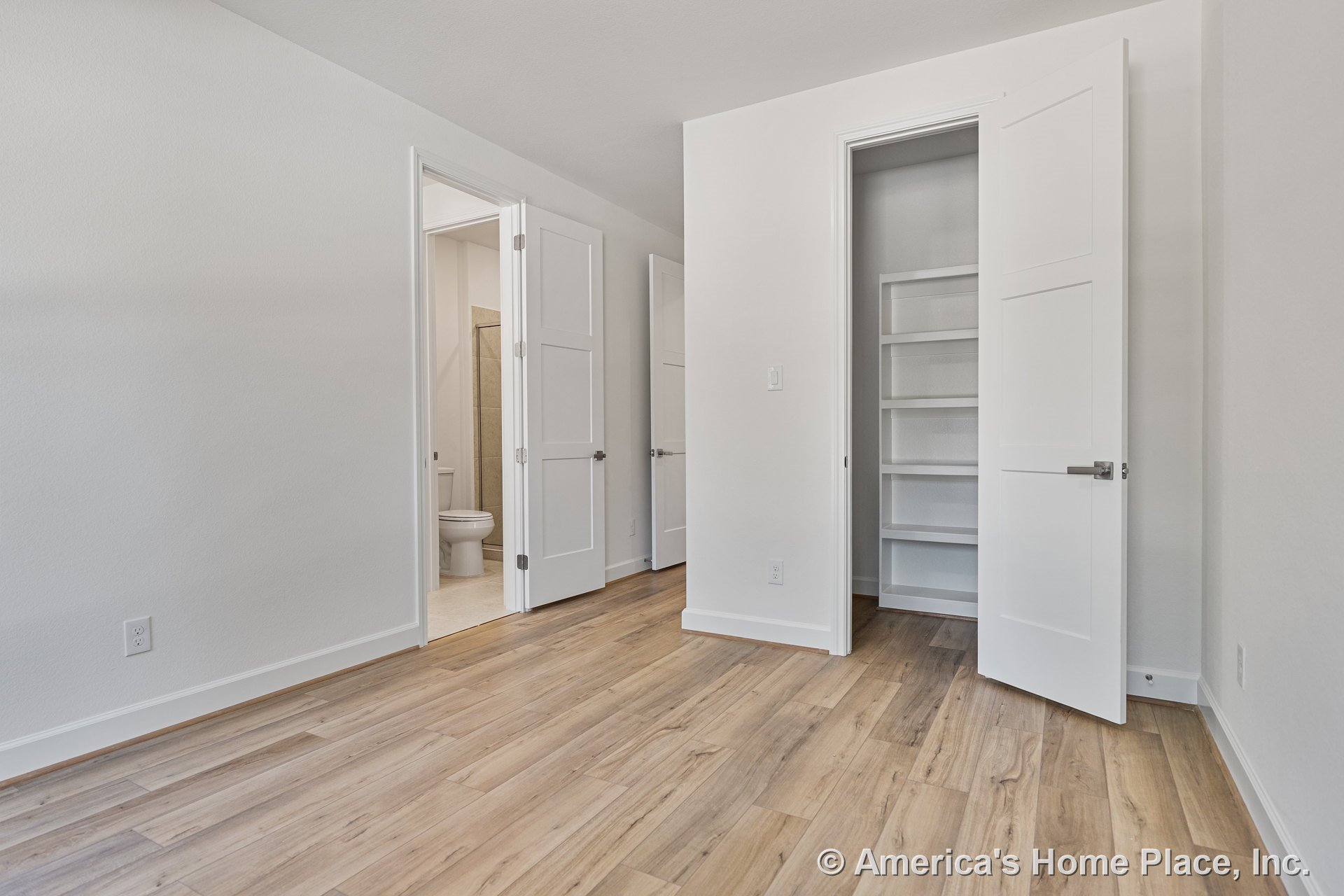 Bedroom with light wood plank flooring, white paneled doors, built-in closet shelving, baseboard trim, and entry to en-suite bathroom; neutral wall paint throughout.