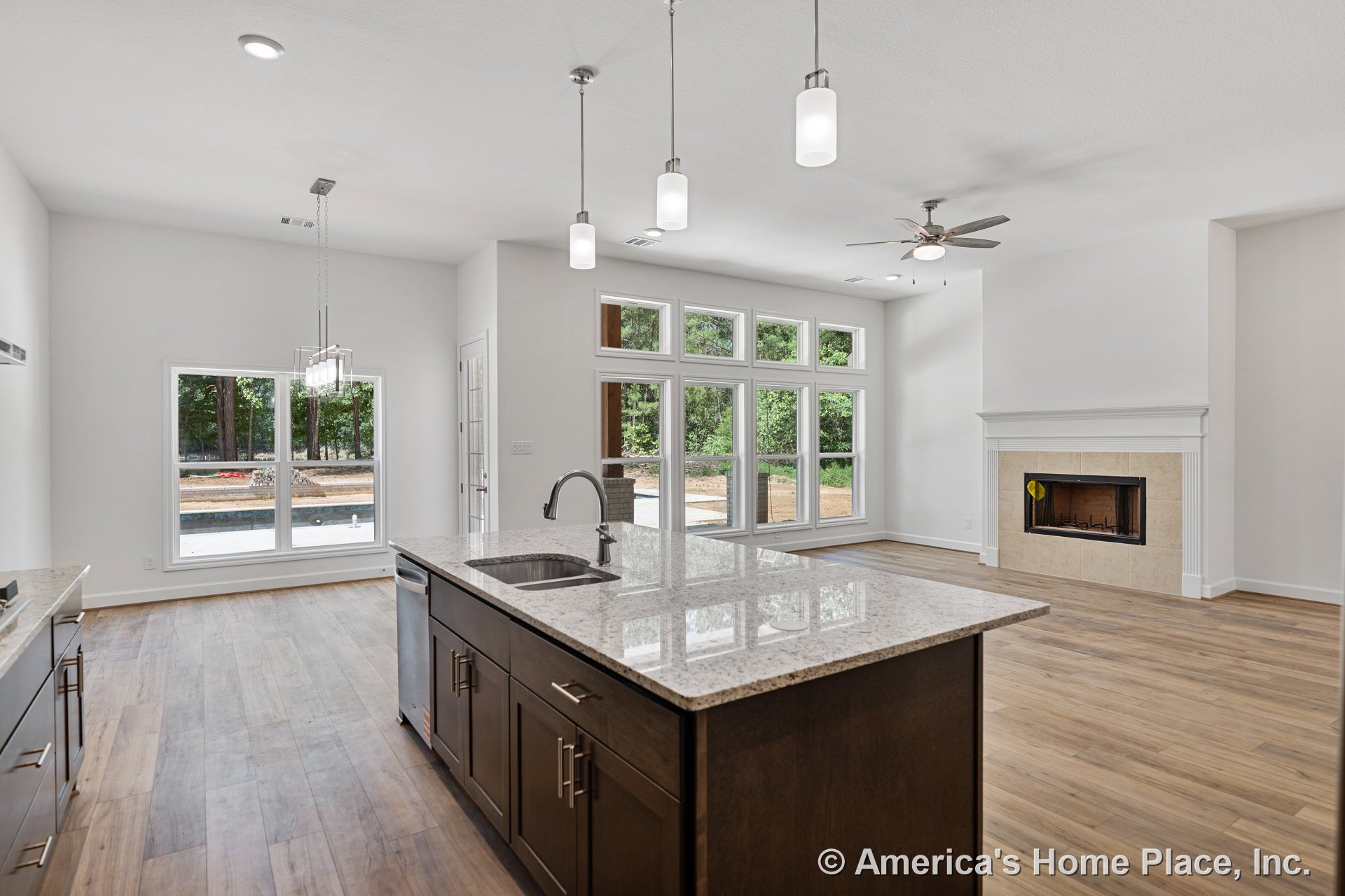 Spacious open kitchen and living area with a large granite island, dark wood cabinetry, pendant lights, wide plank wood flooring, multi-pane windows, tiled fireplace surround