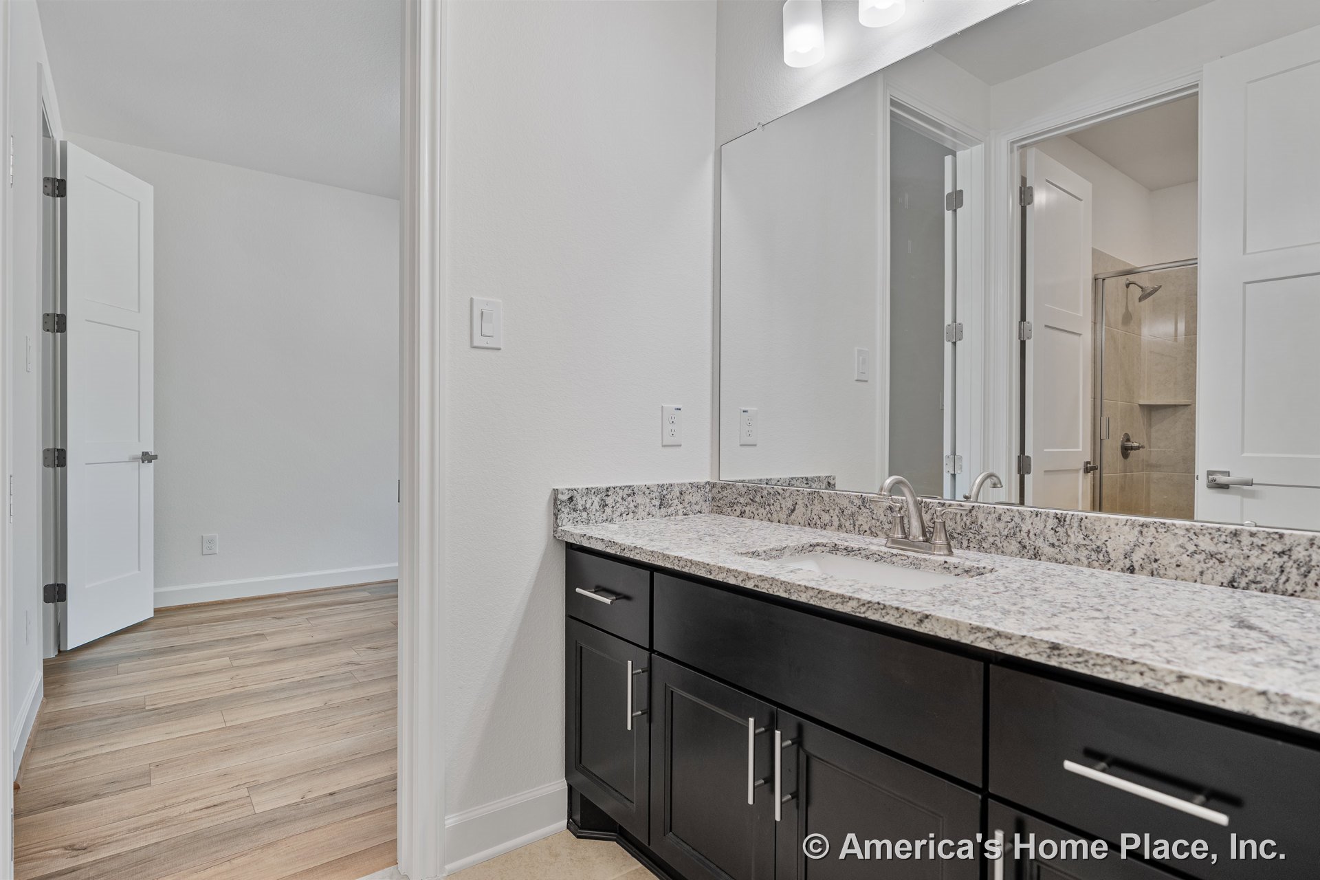 Granite vanity with undermount sink and dark wood cabinetry, large wall mirror above, recessed lighting, tile shower enclosure, white paneled doors, neutral walls, wood flooring