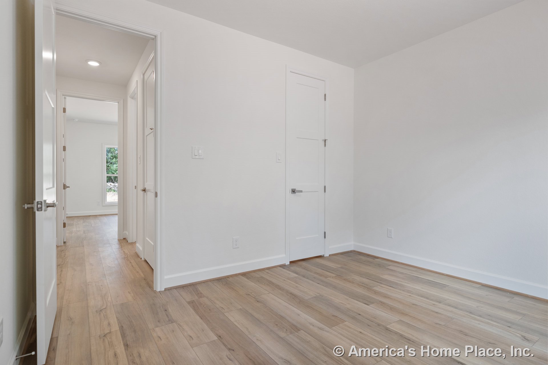 Bedroom with light wood plank flooring, white baseboards, multiple white interior doors, recessed ceiling light, and neutral wall paint; window visible in adjacent room.