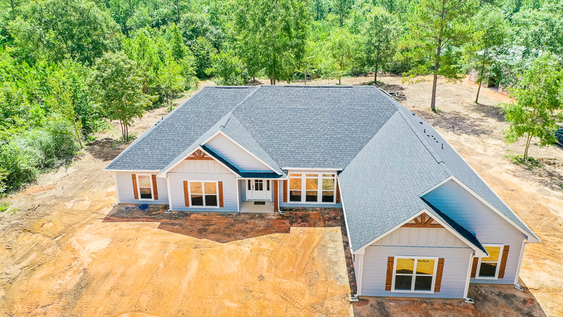 Wood-framed house under construction with exposed roof trusses, surrounded by mature trees and dirt ground