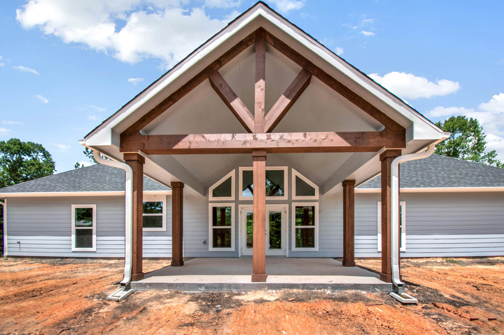 Partially built house with expansive roof, white-framed window, unfinished porch area, dirt patch, and blue sky with scattered clouds