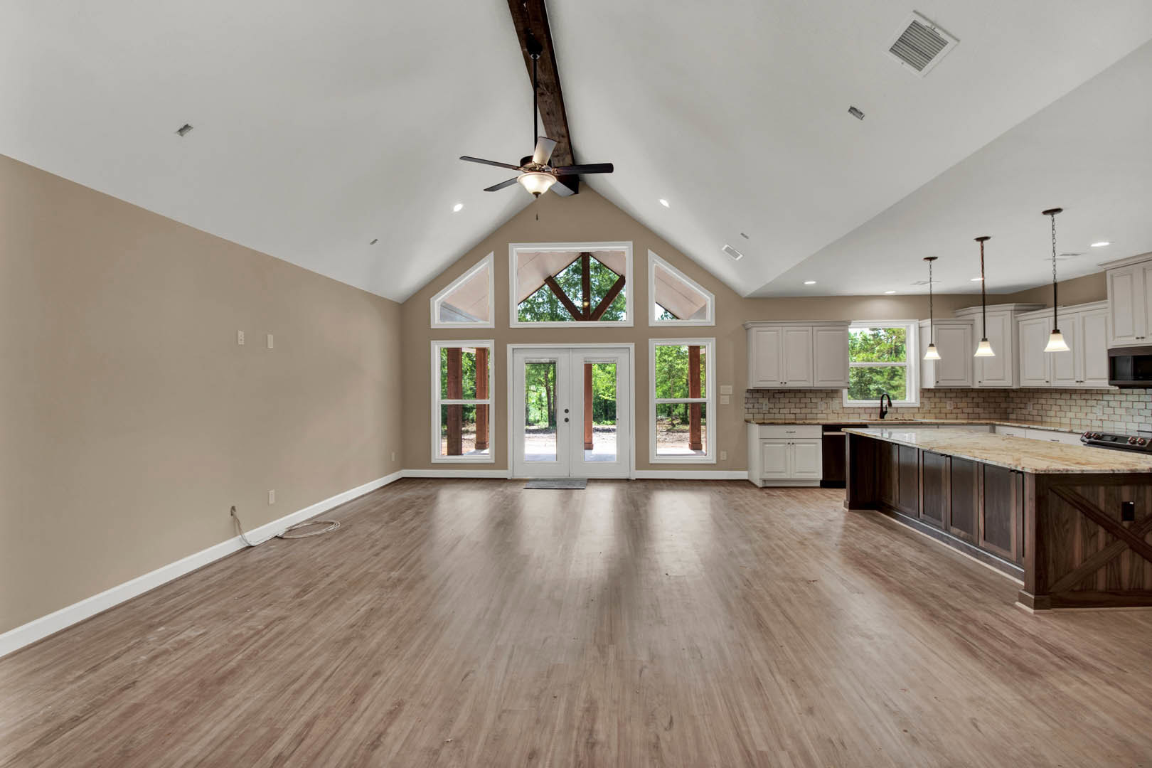 Spacious open floor plan featuring hardwood flooring, ceiling fan with light fixture, triangular window, double glass-paneled doors, white-framed accents, and visible kitchen