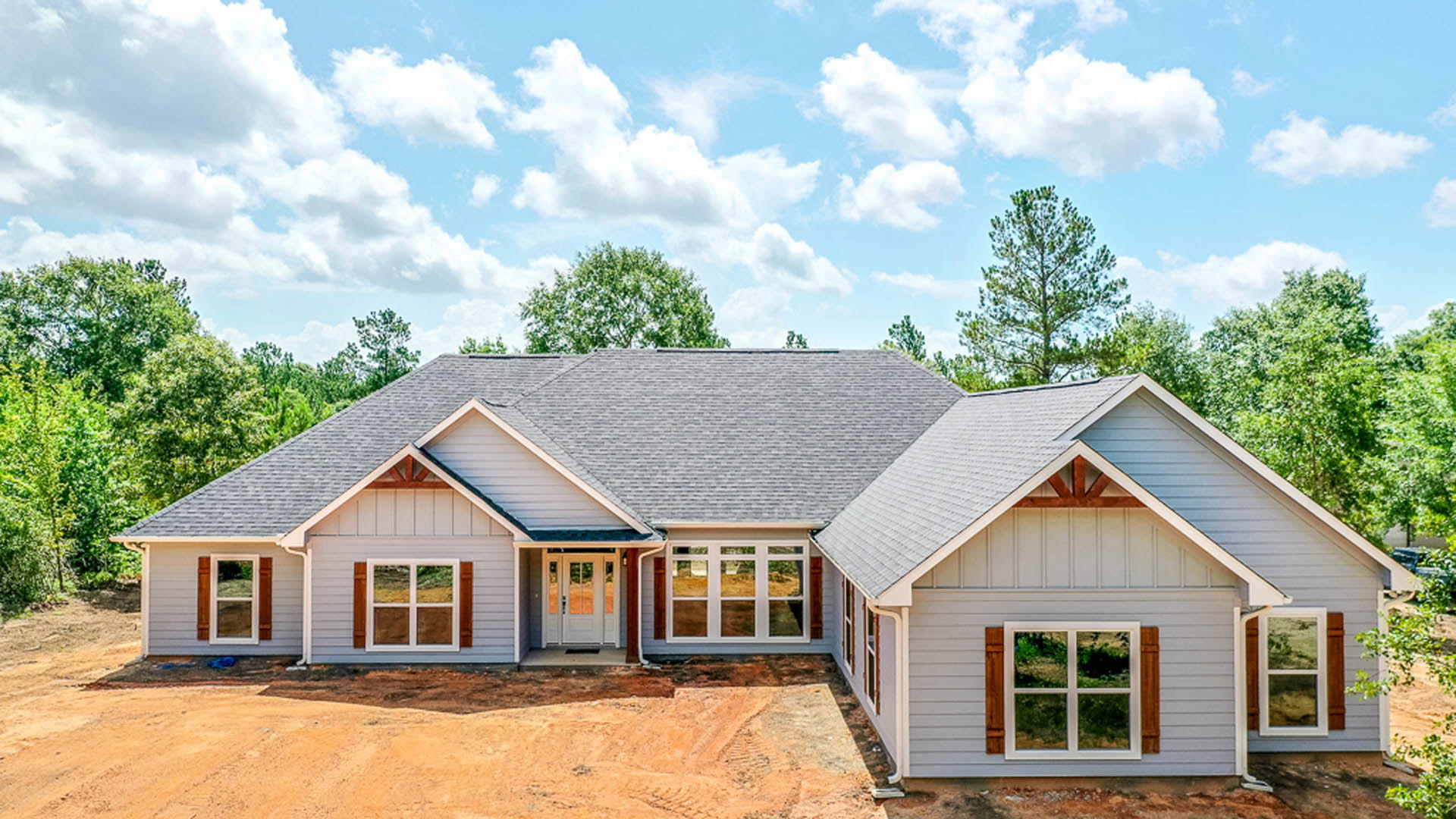 Partially built house with exposed framing, large white-framed windows, glass-paneled front door, surrounded by trees under a blue sky with scattered clouds