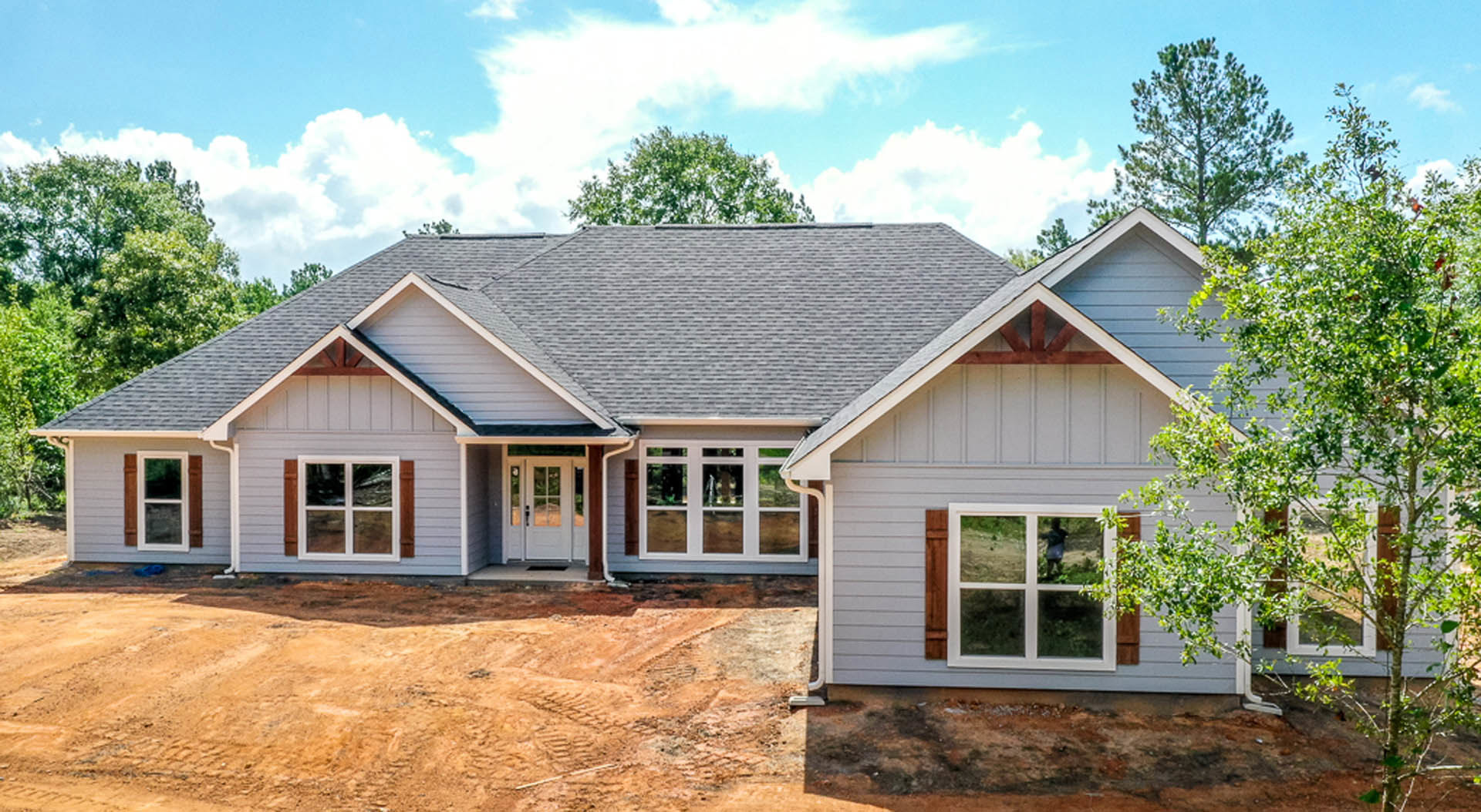 Framed house under construction with exposed plywood, large dirt driveway, scattered construction materials, and mature trees in the background