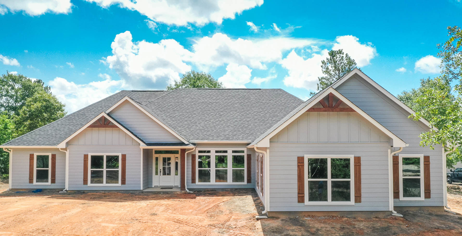 Wood-framed house under construction with white-trimmed windows, glass-paneled front door, and pitched roof, set against a clear blue sky and leafy tree.