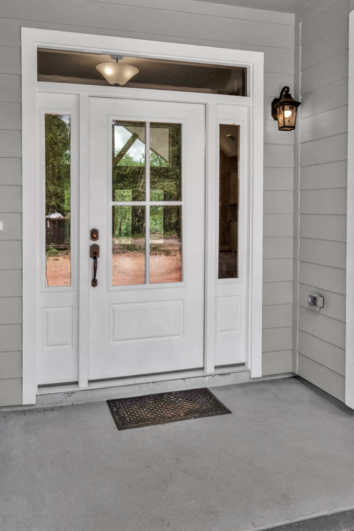 White paneled door with glass panes, metal grate detail, window overlooking forest, illuminated light fixture, textured floor mat, hardwood flooring