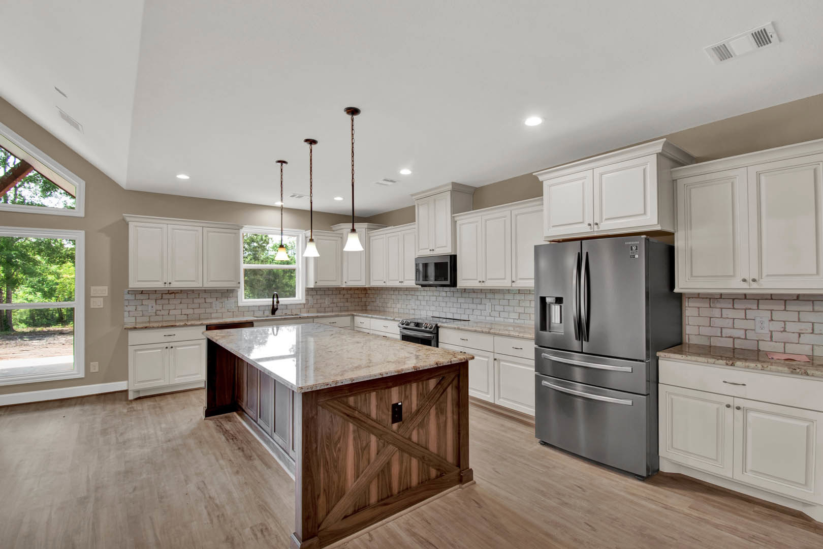 Spacious kitchen featuring a marble-topped island, stainless steel refrigerator, glass-door microwave, white cabinetry, and large window overlooking leafy trees