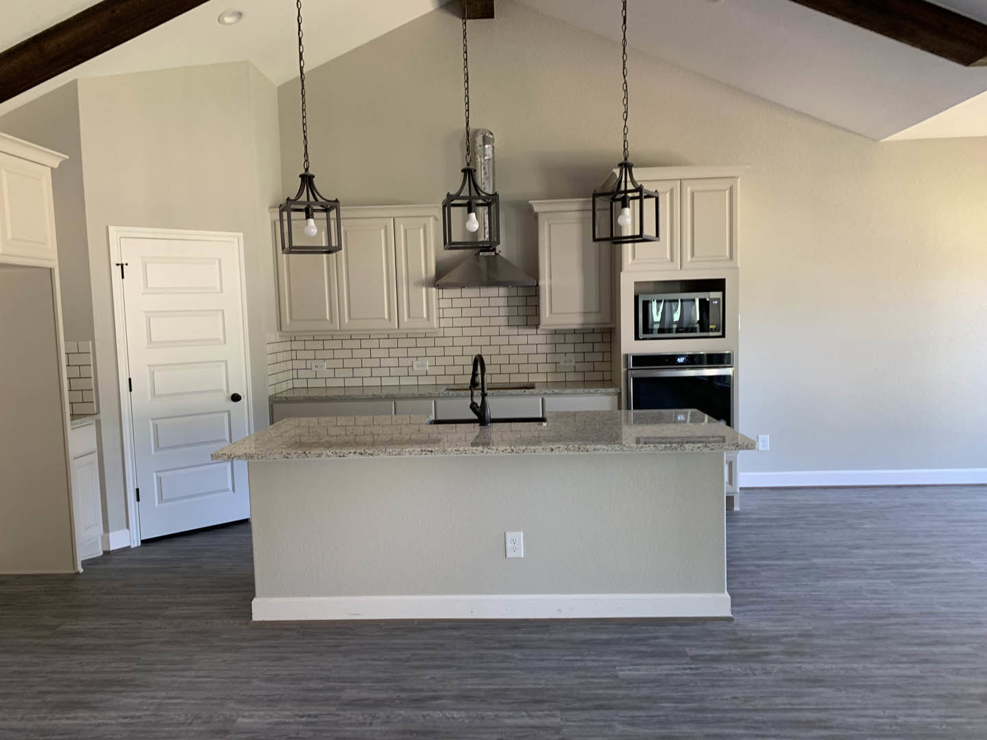 Modern kitchen featuring a central island with built-in stove, white walls, black sink and faucet, grey flooring, white door with black knob, window with sheer curtains, and