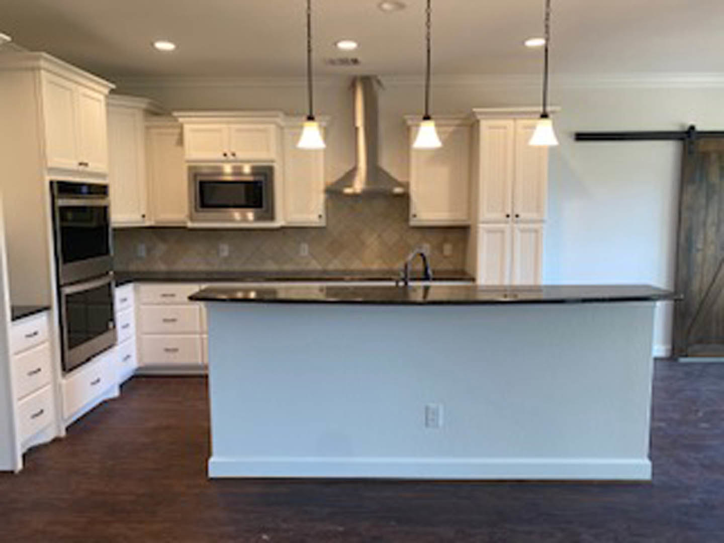 White kitchen cabinets and black countertop with stainless steel sink, white wall, blurry microwave, chair, and window in background, light switch visible on wall.