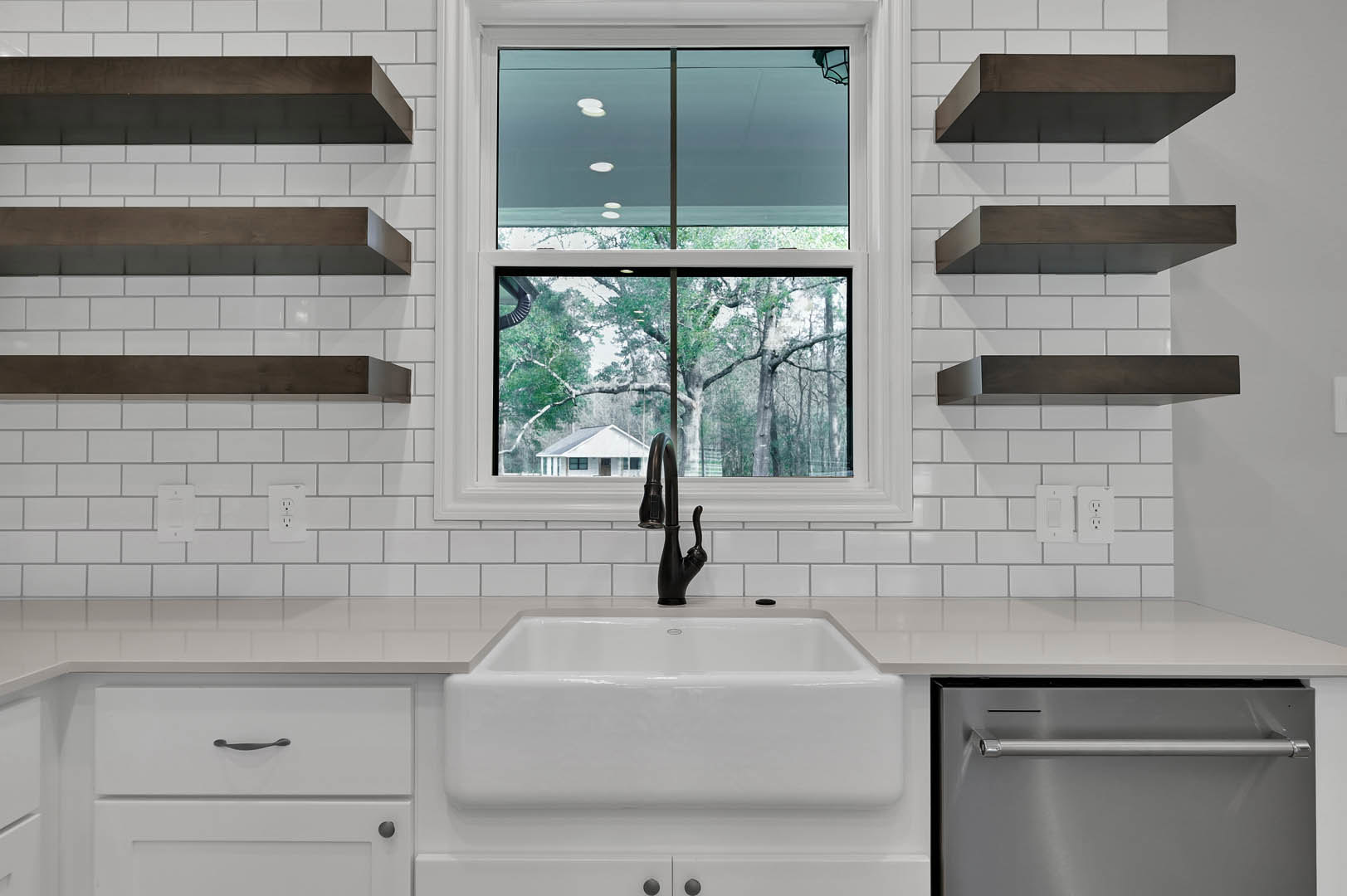 Modern kitchen with white sink, open shelves, silver oven, black countertop appliance, large window overlooking trees and neighboring house