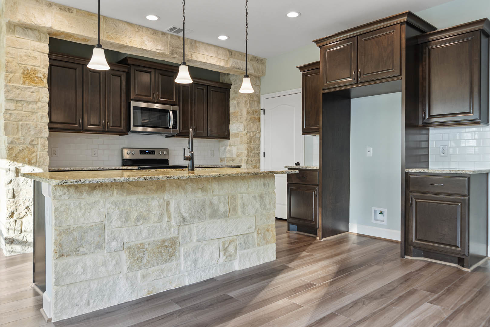 Kitchen featuring textured stone walls, dark wood cabinets, granite countertops, stainless steel microwave, and tile flooring