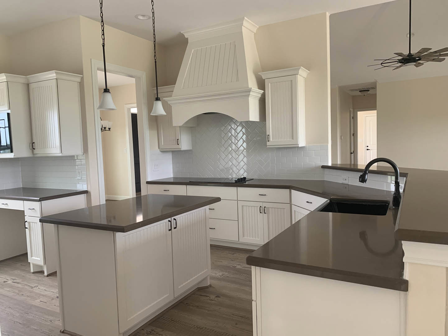 White kitchen with black countertops, white cabinetry, tile backsplash, stainless steel sink and faucet, kitchen island, ceiling fan with light, white paneled door, and modern