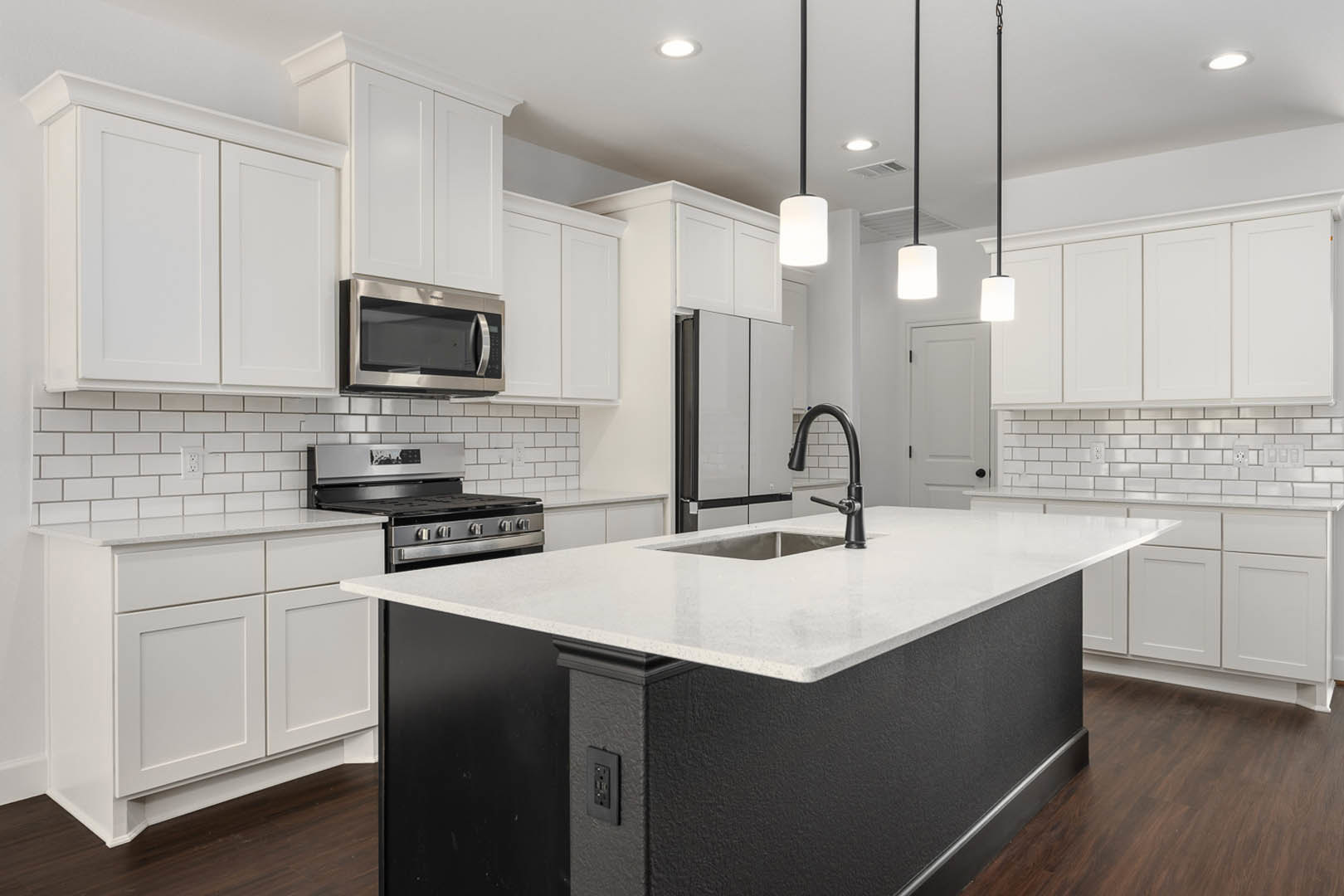 White kitchen cabinets, black and white island with integrated sink, black faucet set against white tile backsplash, built-in microwave, white door featuring black handle, white