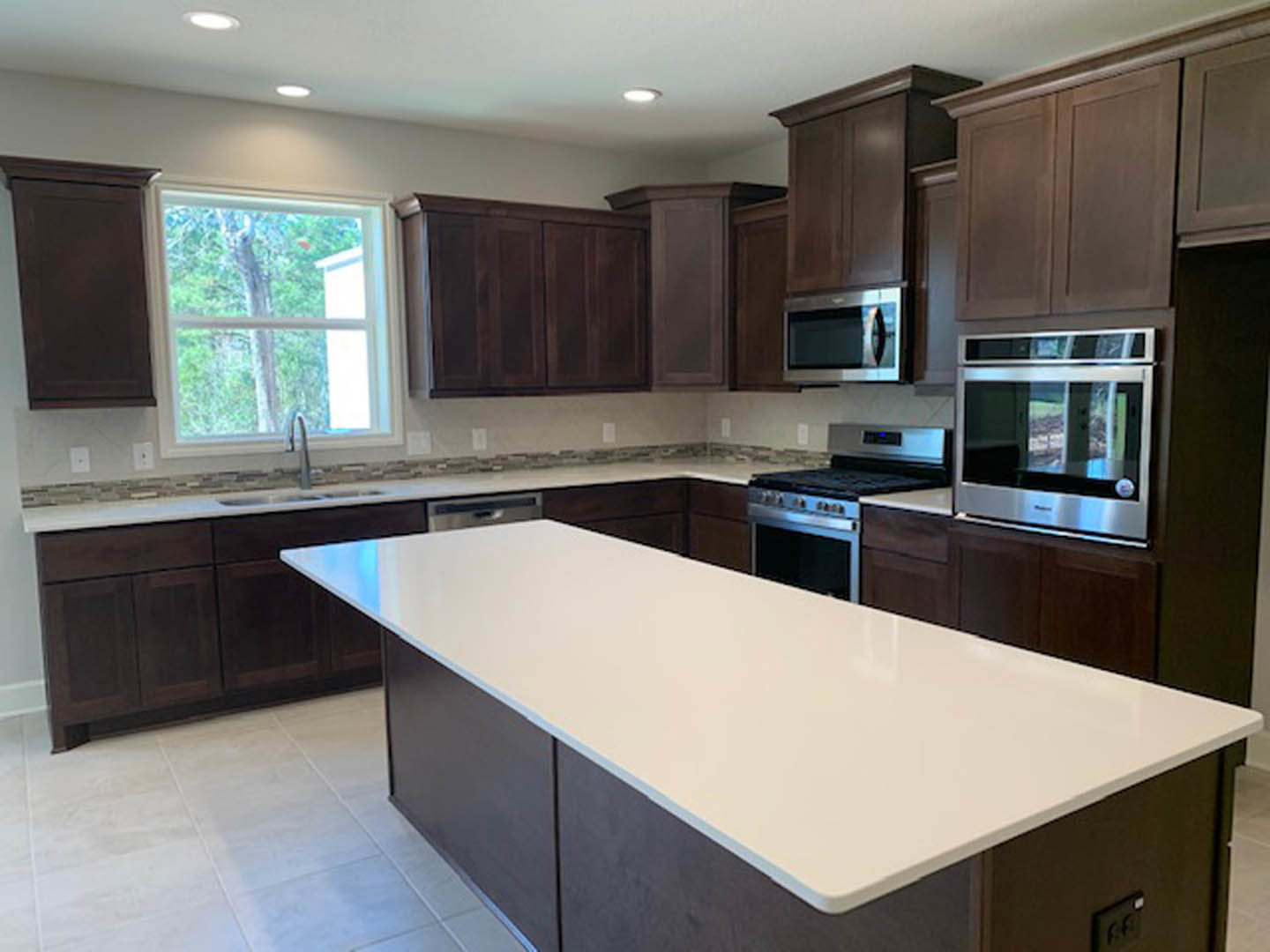 Dark wood kitchen cabinets, white quartz countertop, stainless steel microwave and stove, window overlooking leafy tree, light tile flooring
