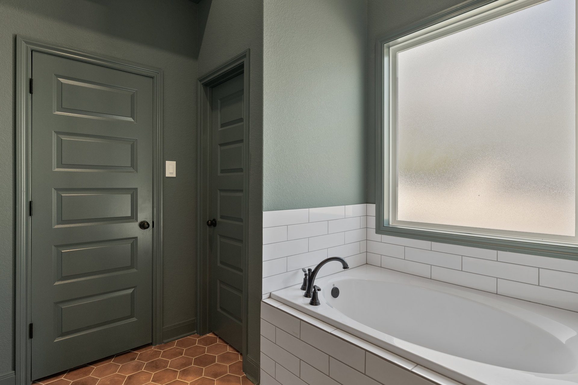 White bathtub with tiled walls, frosted window above tub, black faucet on white sink, grey door with black handle