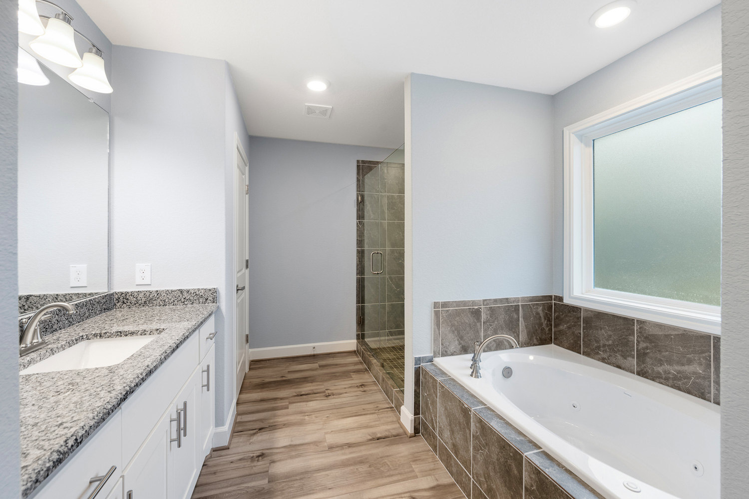 White bathtub set against a tiled wall, adjacent to a modern sink with chrome faucet, under a multi-bulb light fixture, bathroom window visible in background