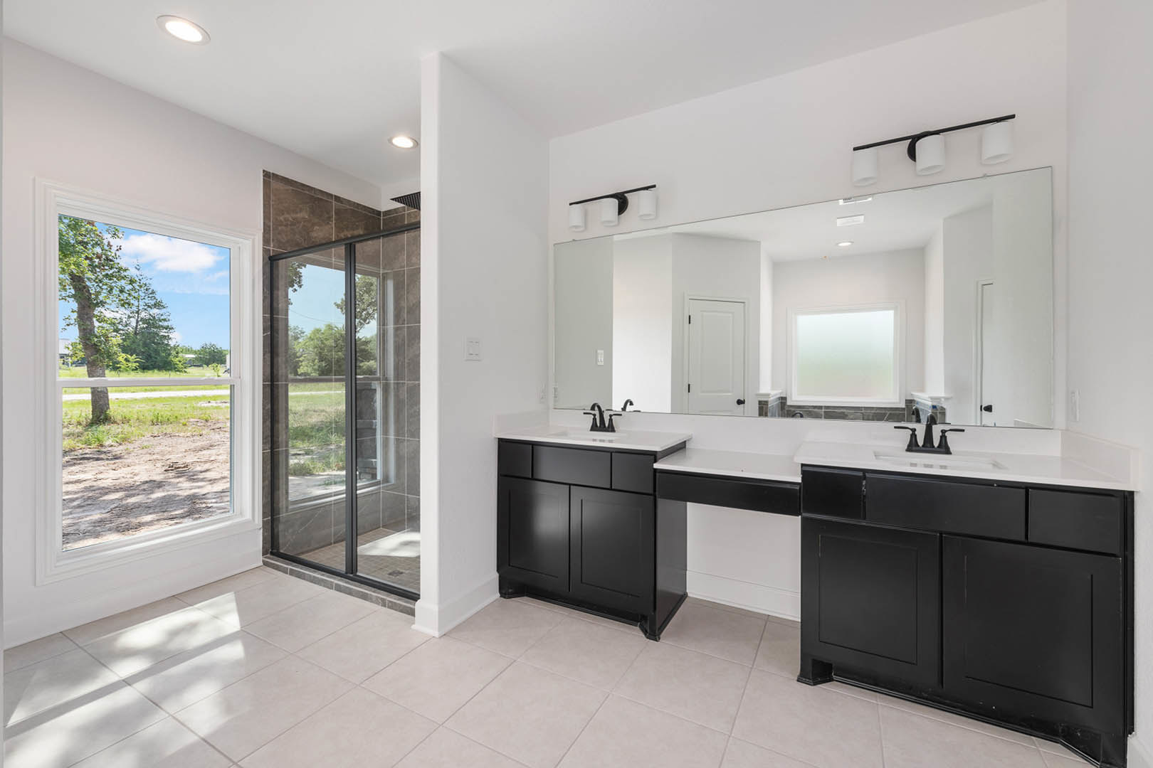 Bathroom with double black sinks on a white countertop, large wall mirror, white cabinetry, tile flooring, and windows showing outdoor views of trees and a field.