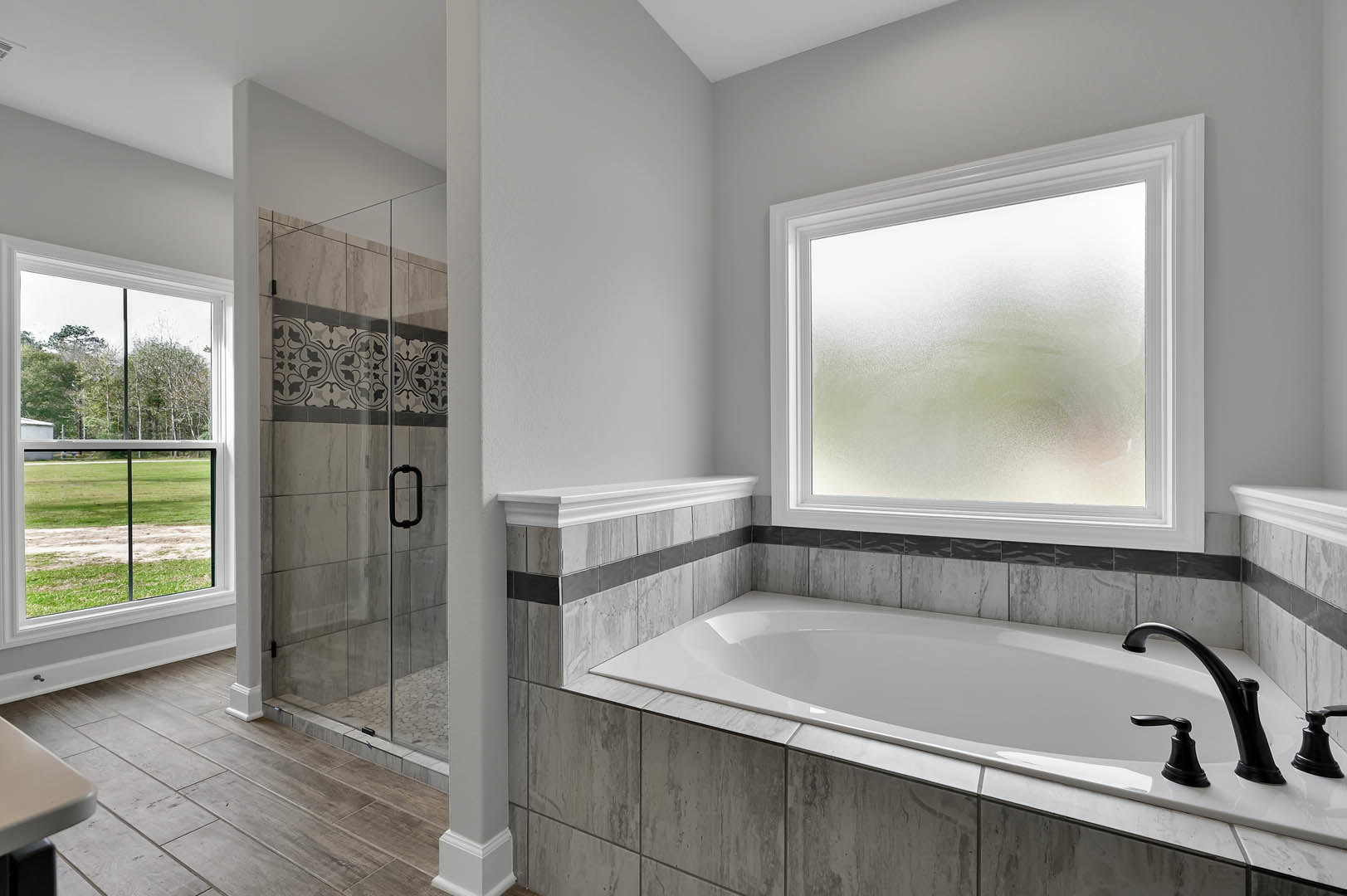 Bathroom with white freestanding tub, glass-enclosed shower, frosted window, black hardware, and light tile walls; window overlooks grassy field.