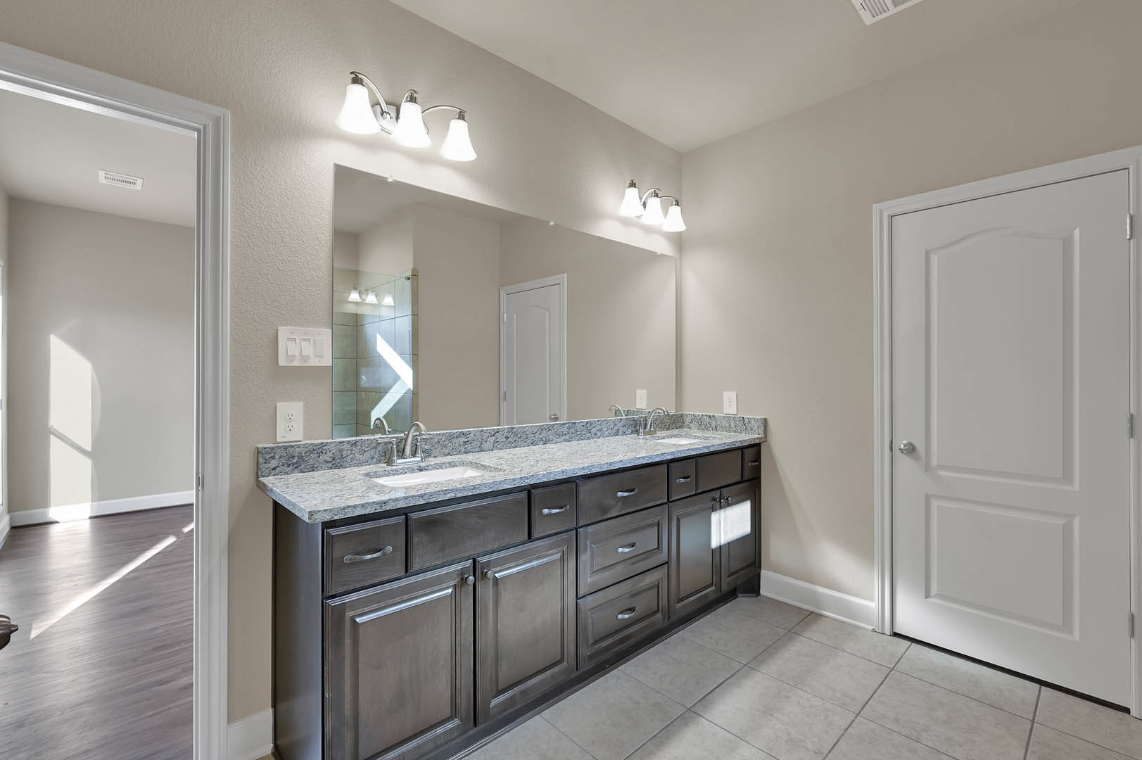 Bathroom with double sink vanity, white cabinetry, stone countertop, silver faucets, tile flooring, white door with silver knob, modern light fixture, three-switch white light