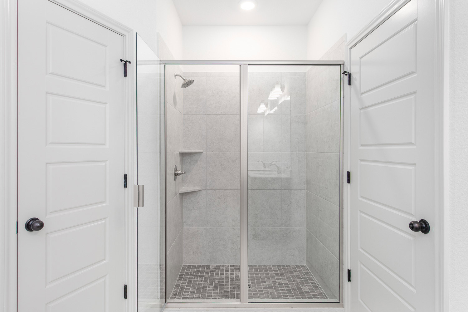 Walk-in shower with clear glass door, chrome shower head, white tile walls, adjacent white door featuring matte black handle