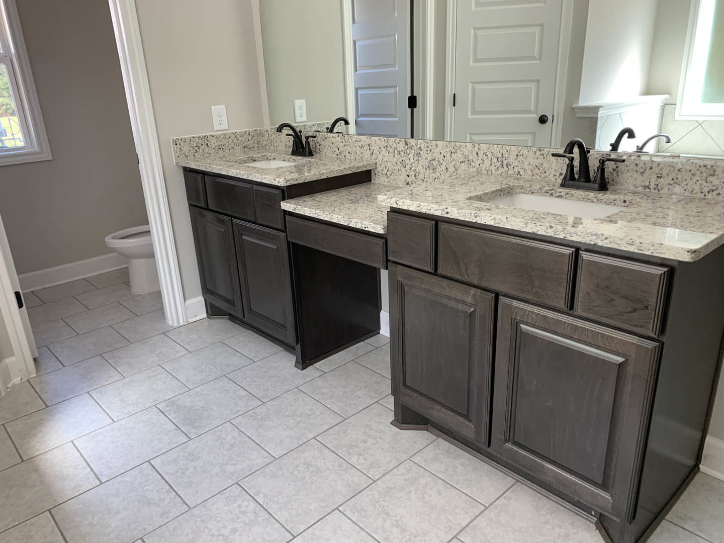Bathroom with marble countertops and dual sinks, white tile floor, black cabinetry, close-up of toilet, window, and white door