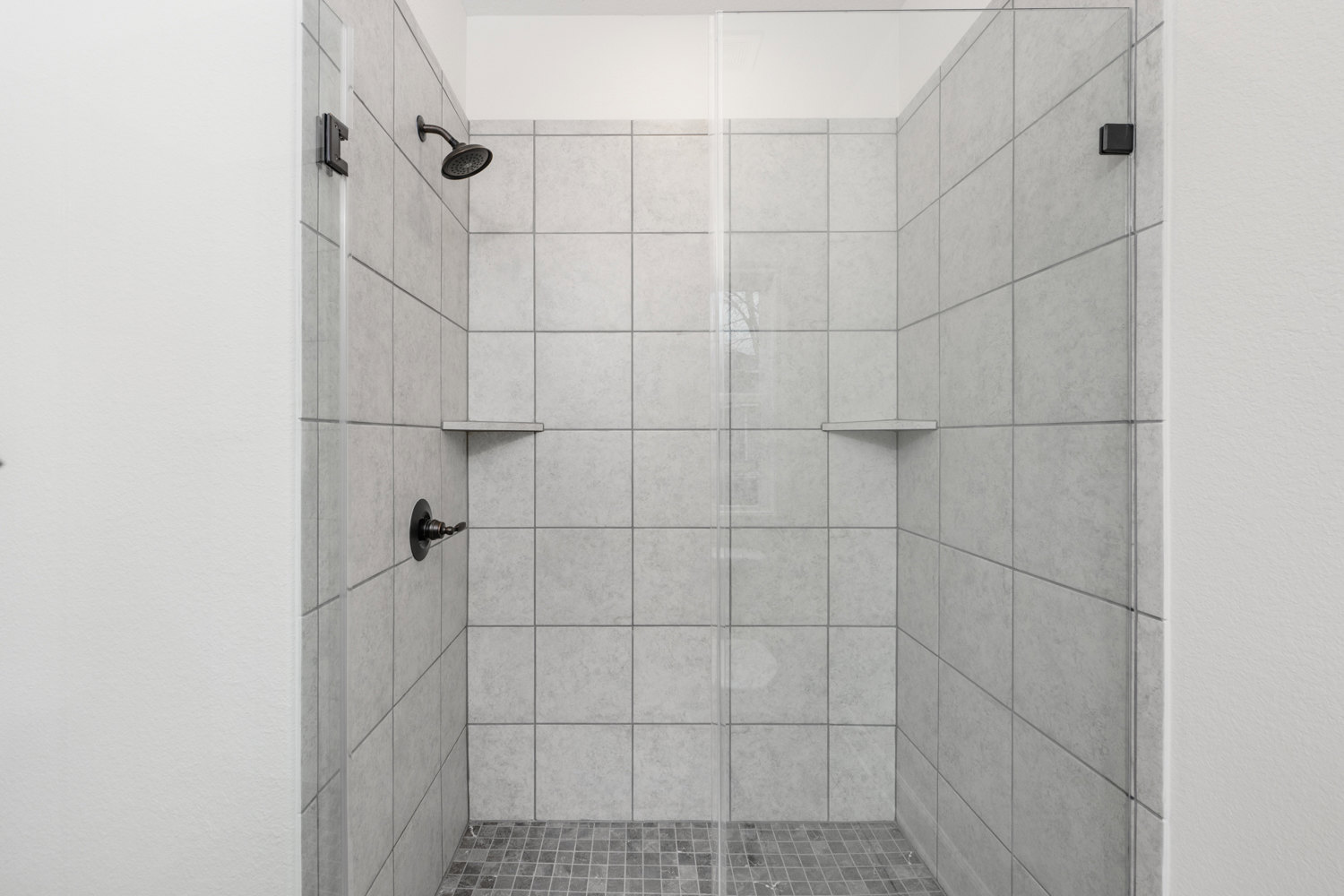Glass shower enclosure with chrome handle, white tile walls and floor, modern shower head mounted on tile wall