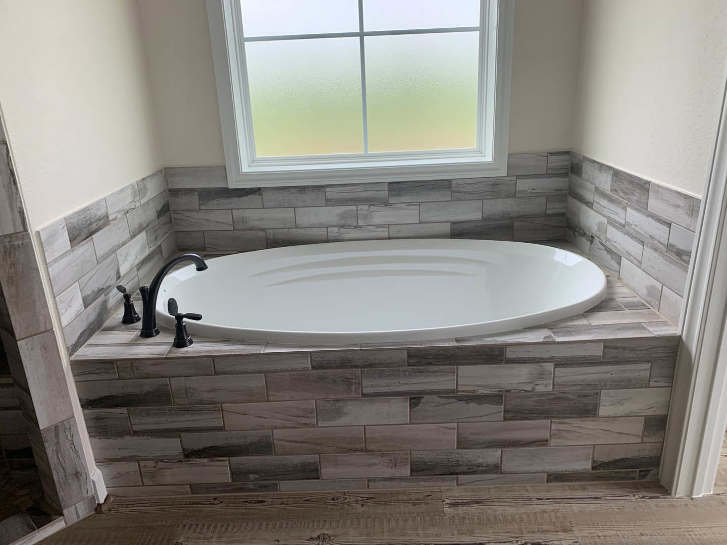 Freestanding white bathtub with chrome faucet beside frosted glass window, surrounded by light tile walls and wood-grained surfaces