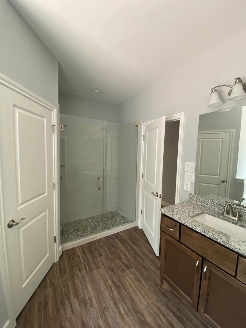 Bathroom with glass shower enclosure, chrome handle, wood flooring, white door, white sink with cabinetry, and dual white lamp light fixture