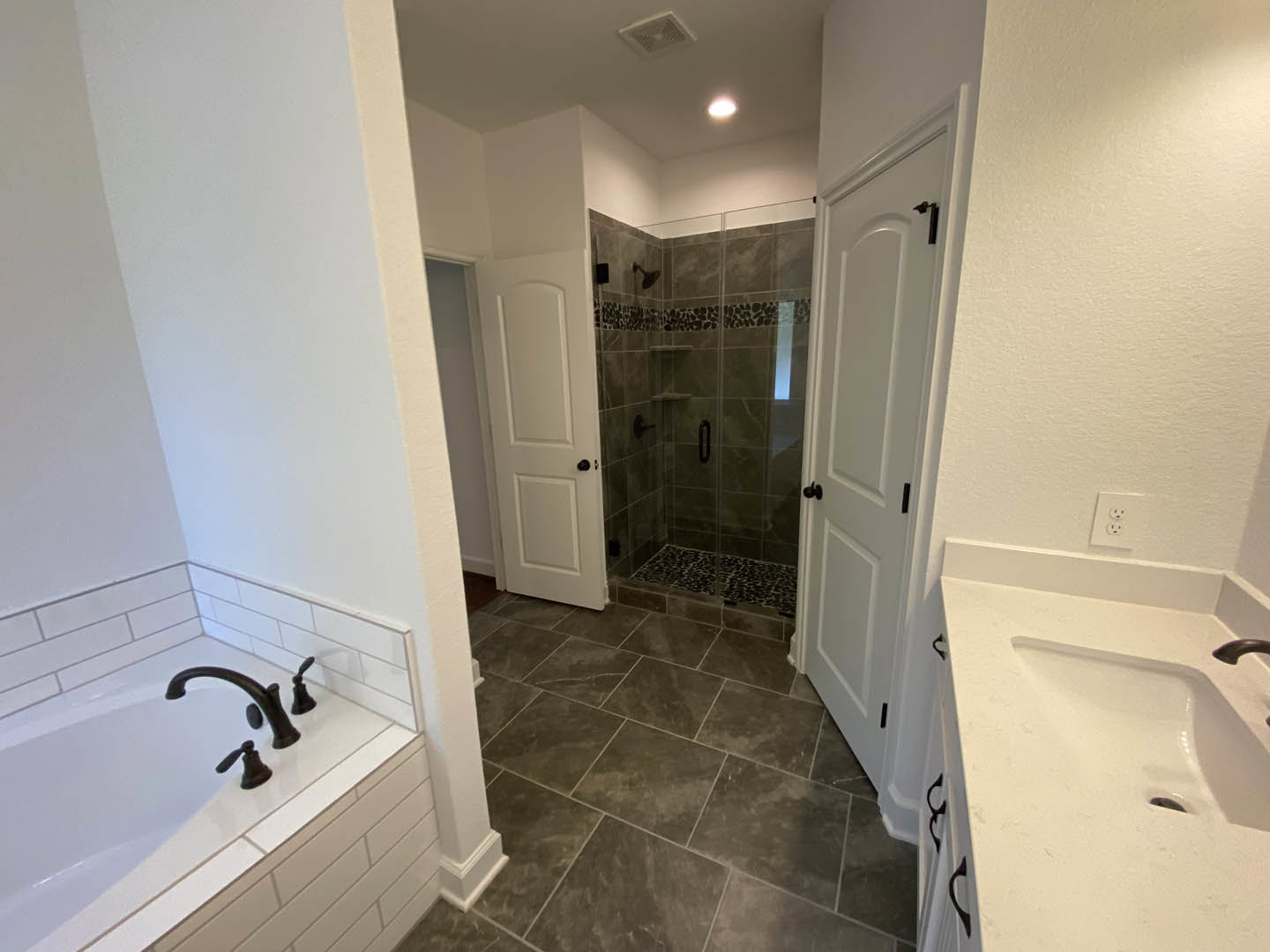 Bathroom with grey tile floor, white sink and bathtub, black faucets, glass shower enclosure, white doors with black handles, light walls and ceiling.
