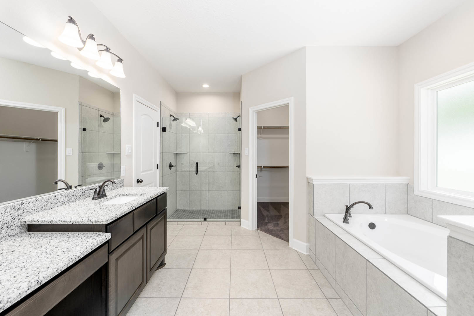 White freestanding bathtub with matte black faucet beside a light wood vanity and undermount sink, glass shower enclosure, white walls, and black hardware on door and window frame