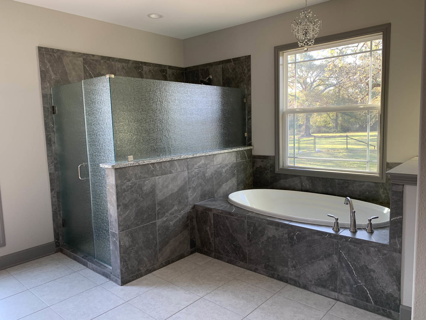 Bathroom with freestanding tub, glass-enclosed shower, white oval sink with silver faucet, white tile floor, crystal chandelier, large window overlooking lawn and trees, wall