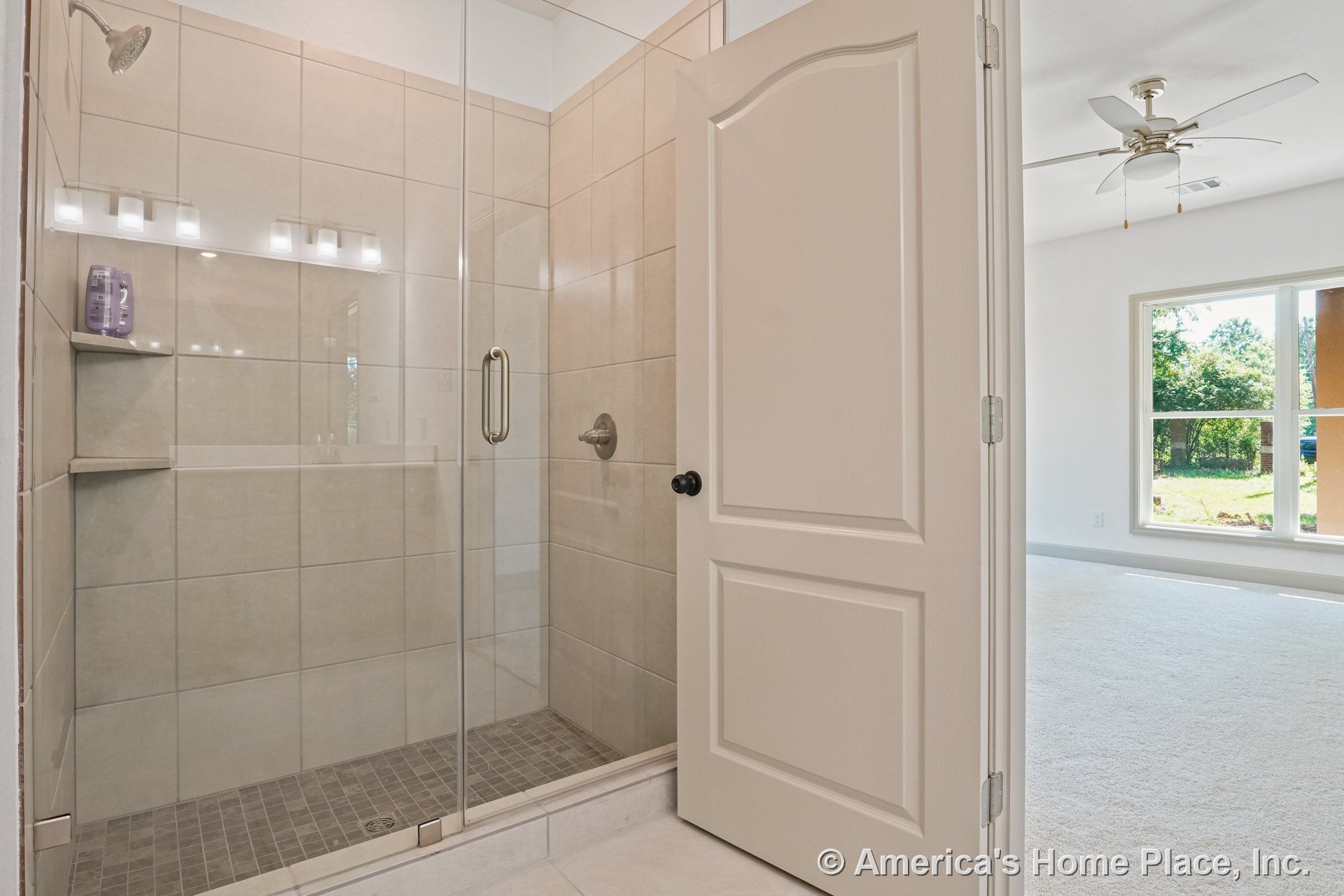 Walk-in shower with large beige tile, glass enclosure, and built-in shelves next to a carpeted bedroom featuring a ceiling fan and expansive double windows.
