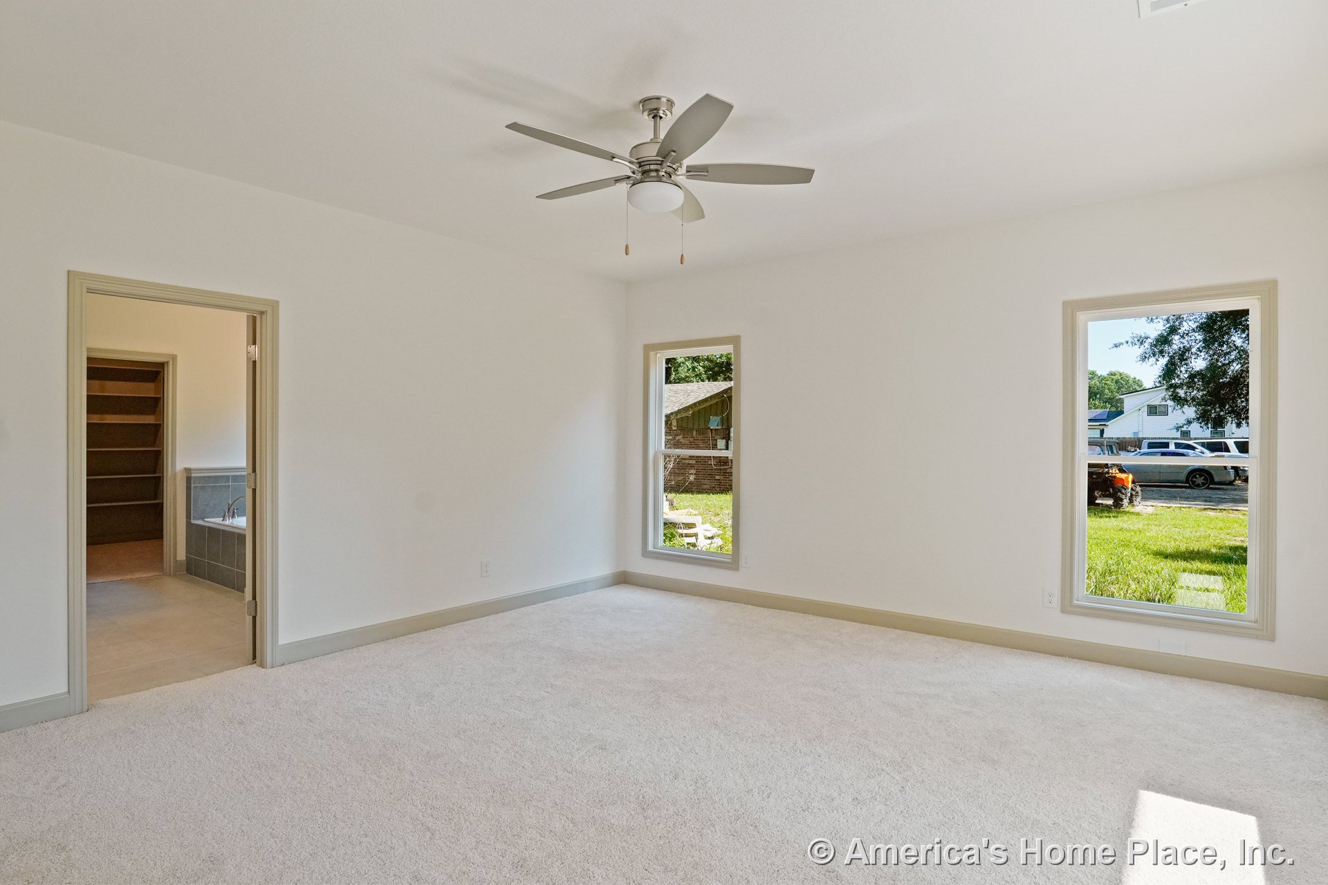 Bedroom with two large windows, light carpet flooring, neutral wall color, ceiling fan with integrated lighting, trimmed doorways, and bathroom entry featuring tile surround.