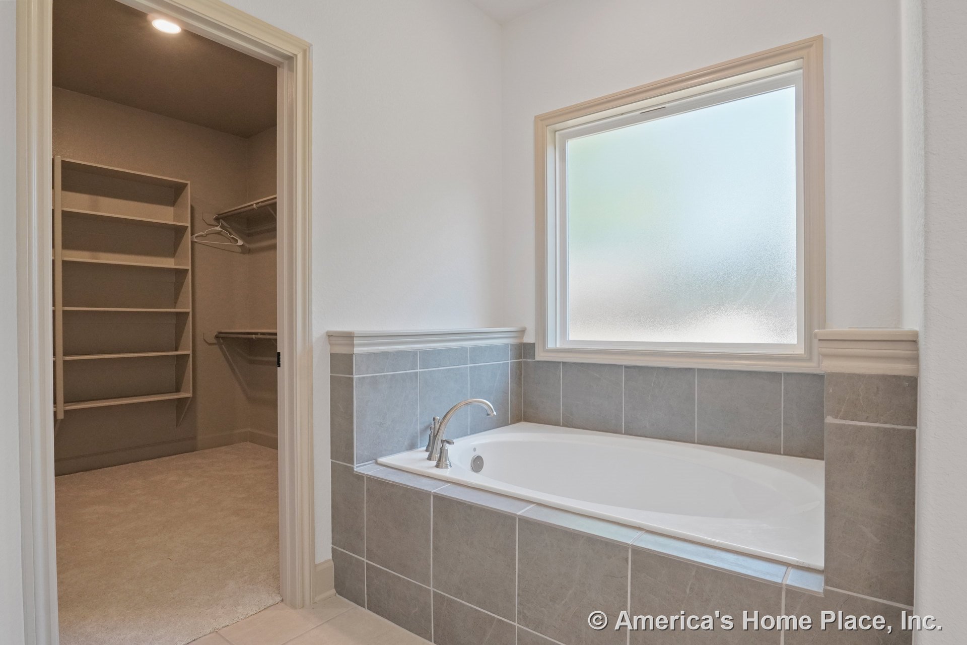 Built-in soaking tub with gray tile surround beneath a frosted window, adjacent walk-in closet featuring built-in shelving and recessed ceiling light; bathroom has neutral wall and