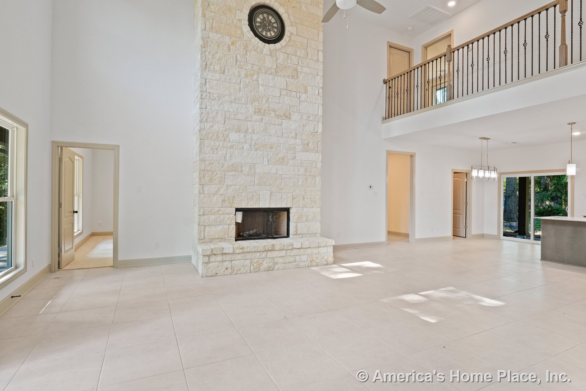 Tall stone fireplace with built-in clock, expansive tile flooring, open balcony with metal railing above, multiple windows with decorative trim, recessed ceiling fan, and modern