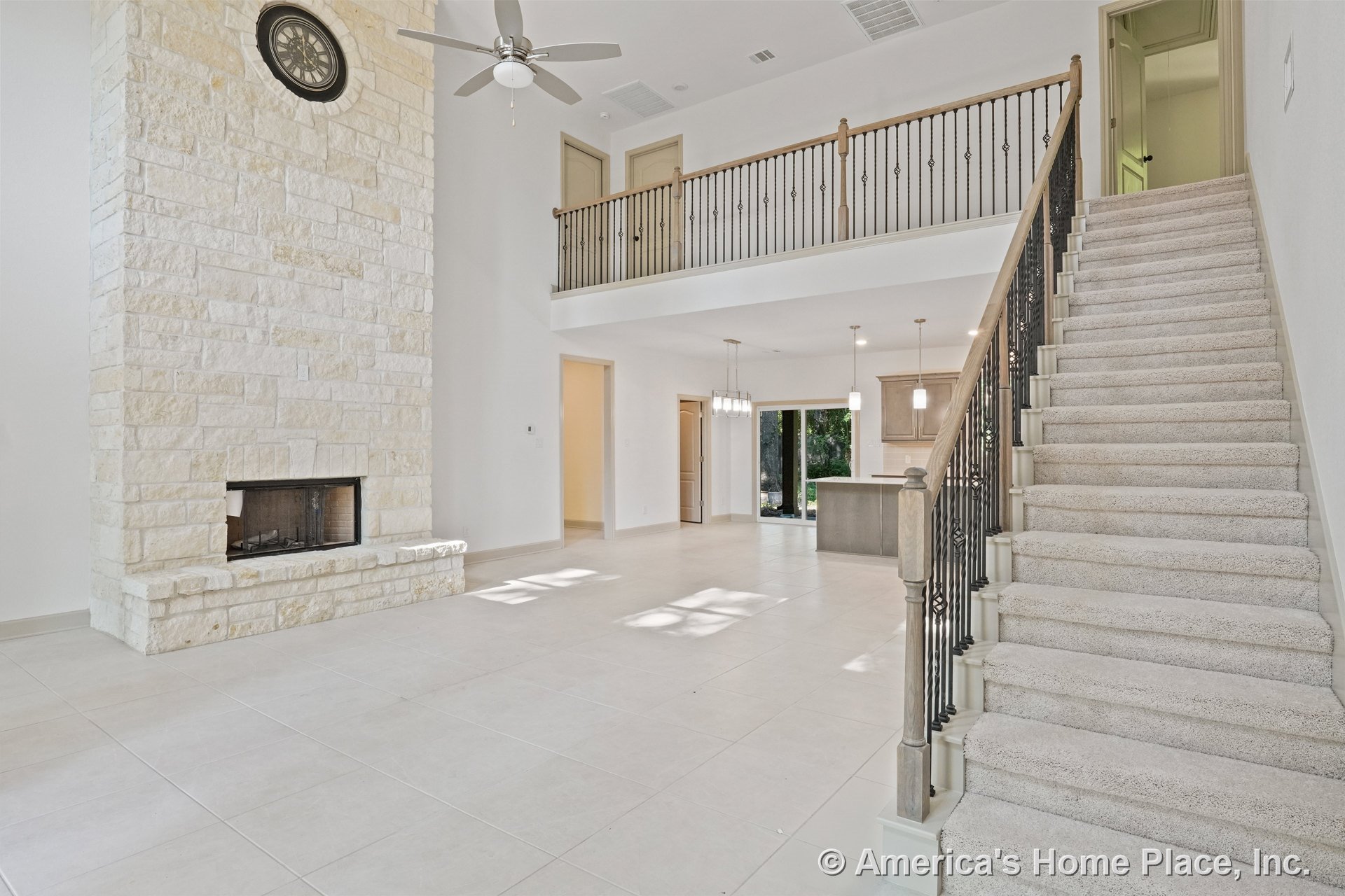 Tall stone fireplace with hearth, expansive tile flooring, open staircase featuring iron balusters, double-height ceiling with ceiling fan, kitchen island illuminated by pendant