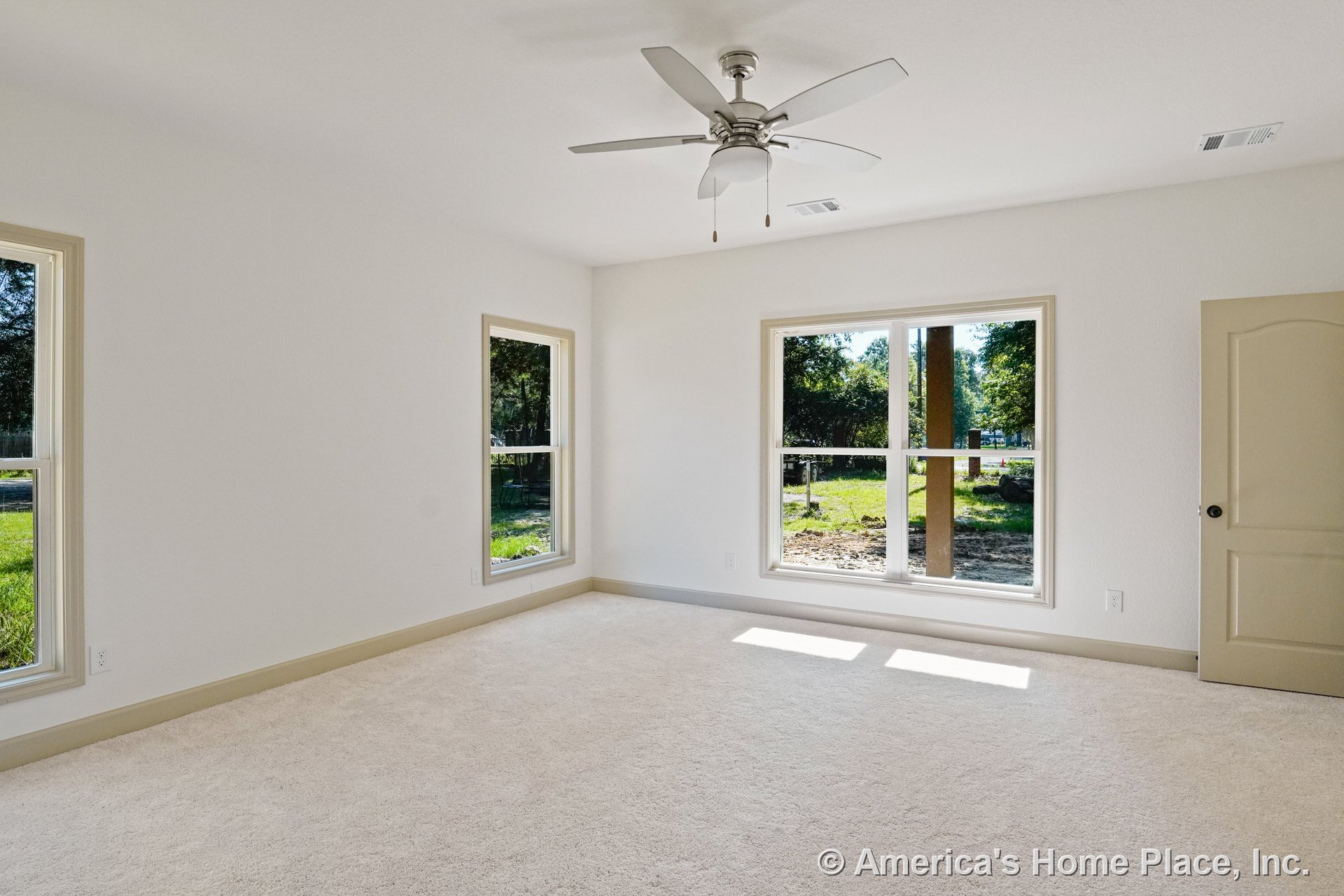 Bedroom with three large windows, neutral carpet flooring, ceiling fan with light, beige trim and baseboards, paneled interior door, and white textured ceiling.
