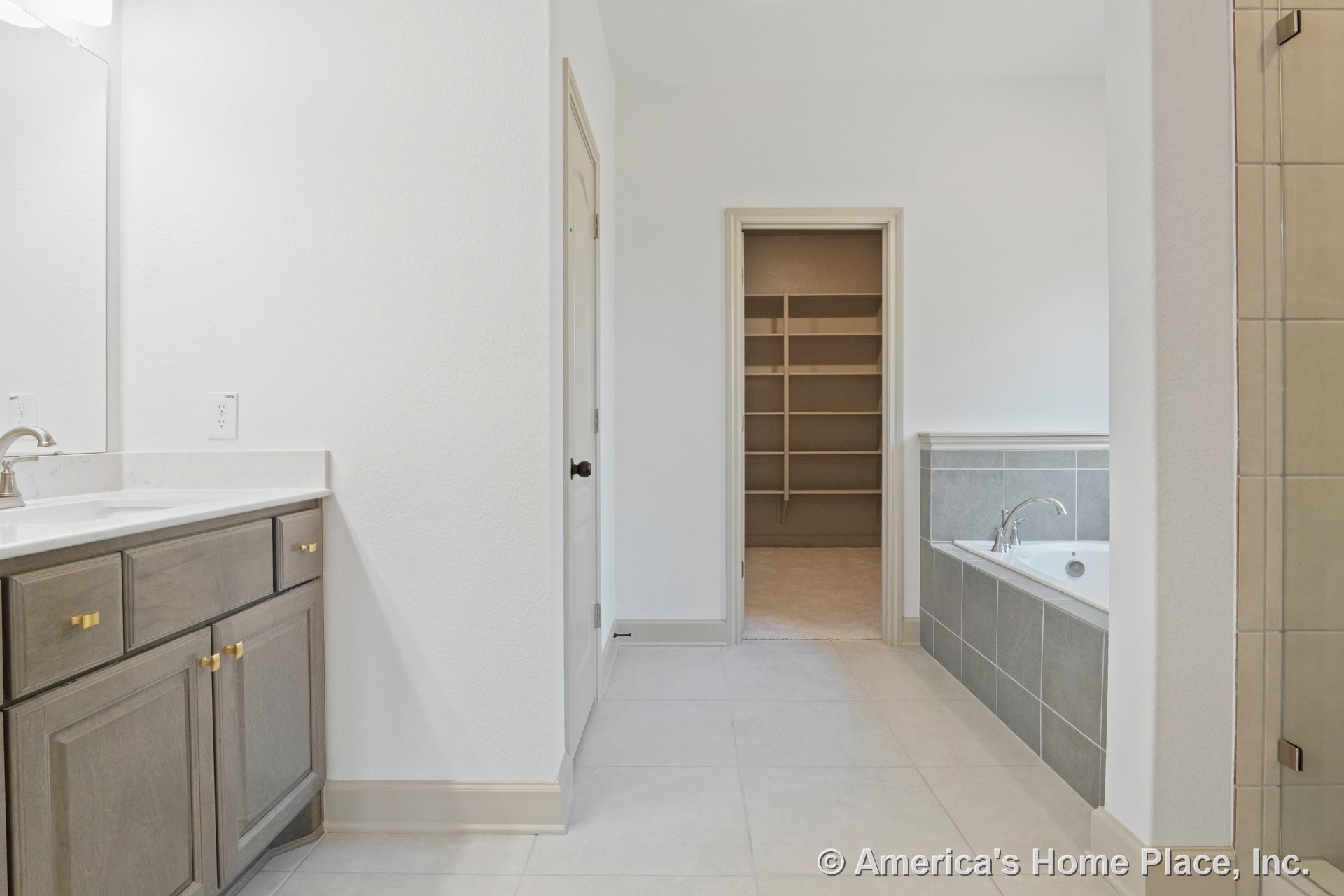 Wood vanity with brass hardware and stone countertop featuring an undermount sink, large wall mirror above, gray tile bathtub surround, walk-in closet with built-in shelving, and