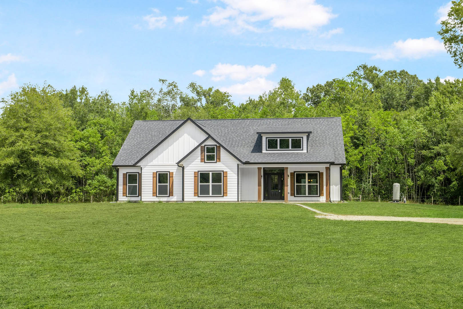 White farmhouse with black roof, white-framed windows, and central white door, set on a green lawn with mature trees and blue sky in the background