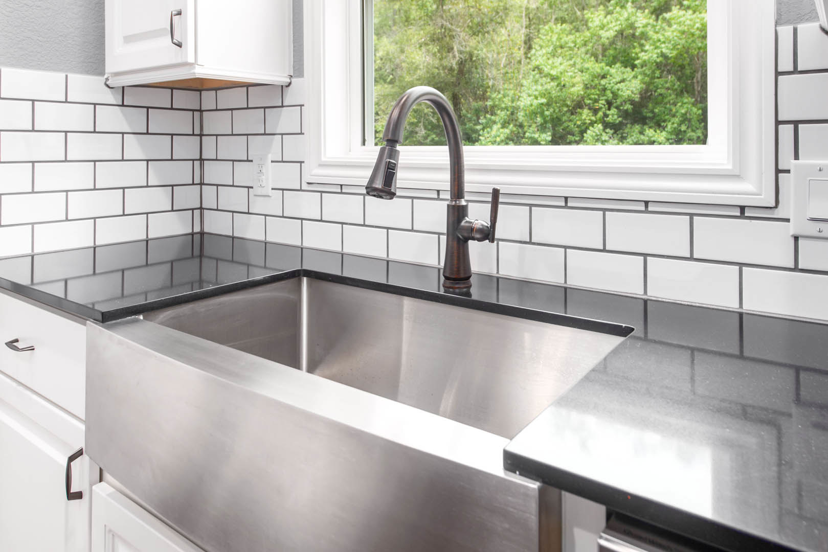 Stainless steel sink with black countertop, white cabinetry, black-handled faucet, tile backsplash, and white electrical outlet in modern kitchen.